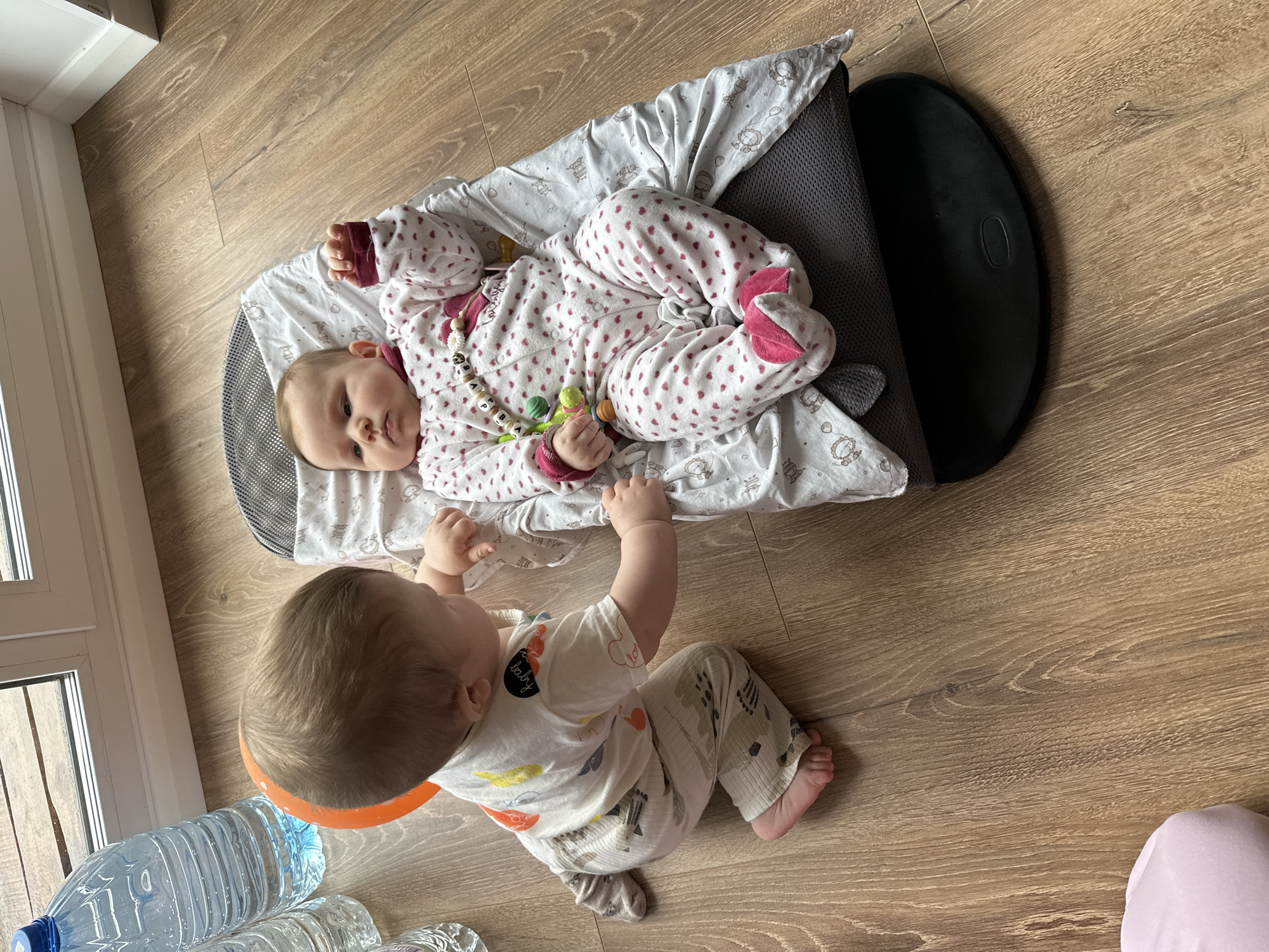 A high-angle, full shot photograph depicts two infants indoors on a wood-patterned floor. One infant is lying in a baby lounger with a grey frame and a patterned white fabric covering. The infant is wearing a white patterned onesie with pink accents and holds a toy. The other infant is sitting upright on the floor, wearing a white patterned pajama set. Several water bottles are visible near the bottom left corner of the frame. The photograph was taken in Chaikovsky, Russia.