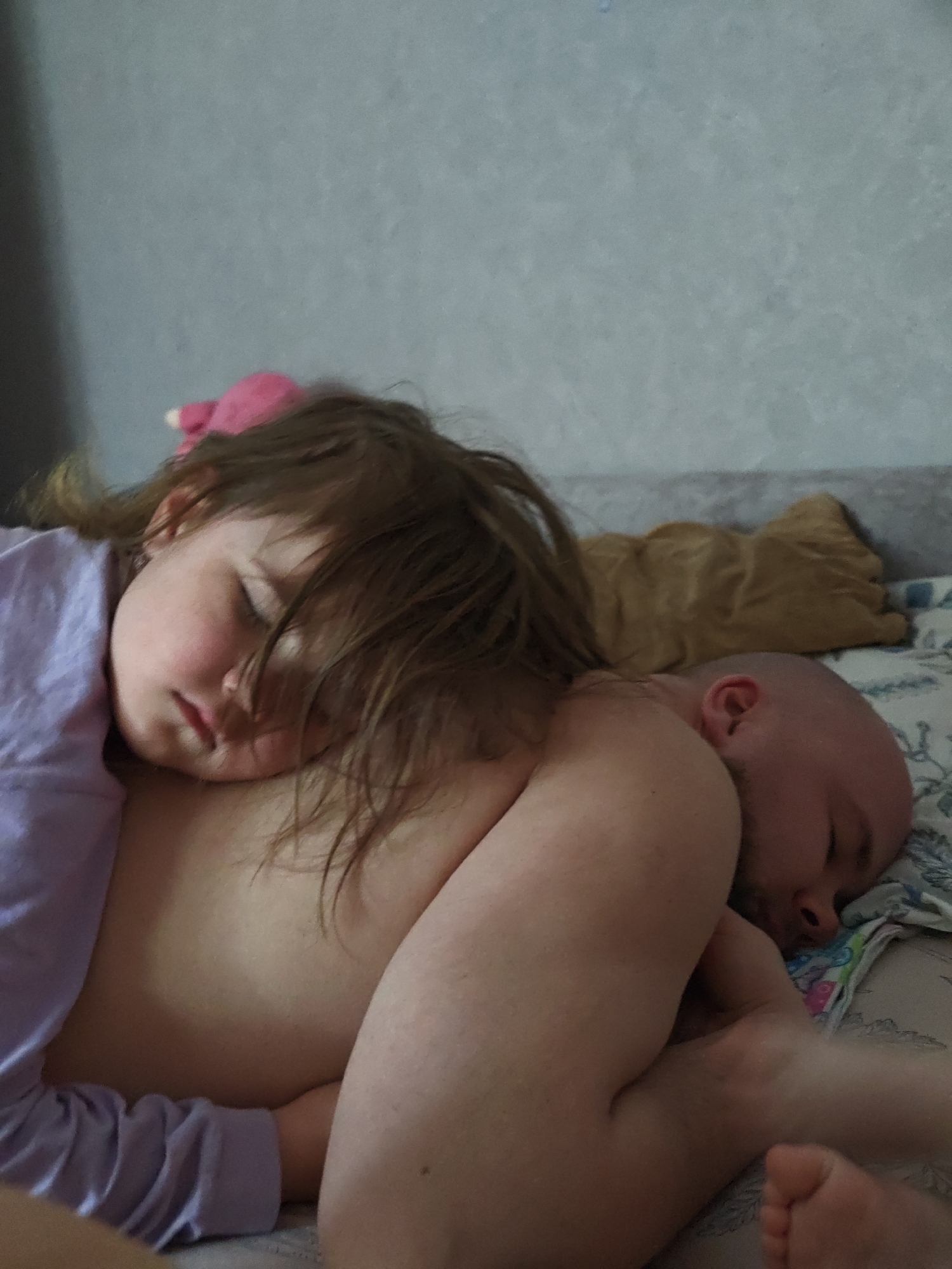 A photograph shows a child and a bald adult sleeping on a bed. The child, wearing a lavender long-sleeved shirt, is resting their head on the adult's chest. The adult is lying on their side. A stuffed animal is in the background, resting on the bed. The bedding has a patterned design. The wall behind the bed is a light color.
The location is not discernible from the image. The claim of the location being Chaikovsky, Russia cannot be supported by the visual evidence in the photograph.