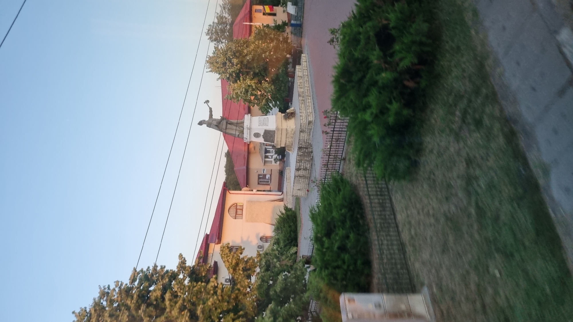 This image depicts a monument in the town of Călugăreni, Romania. The monument features a bronze statue of a man holding a mace, standing on a stone pedestal. The pedestal is adorned with plaques and a wreath of flowers. The monument is situated in a paved square, surrounded by manicured bushes and trees. In the background, two buildings with red roofs are visible, one of which appears to be a public institution with a Romanian flag displayed. The sky is clear and blue, suggesting it is daytime. The perspective of the photo, taken from a slightly elevated and moving viewpoint, suggests it was captured from a vehicle.