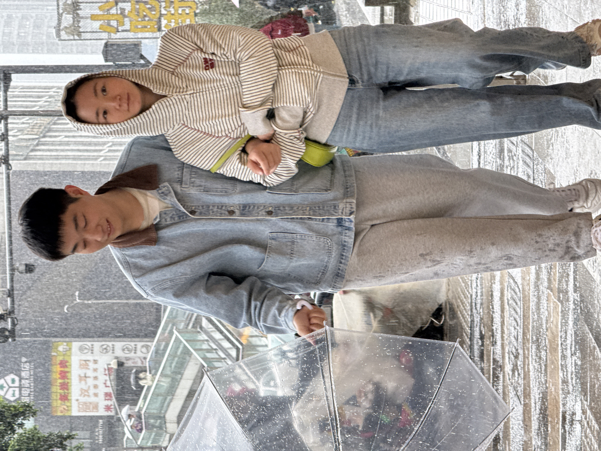 Two people are standing under an umbrella in the rain. The person in the foreground is holding the umbrella and is wearing a light blue denim jacket and grey sweatpants. The person behind them is wearing a striped hoodie, jeans, and white sneakers. The location appears to be an urban environment in Jinjiang District, China, based on the presence of Chinese signage. Rain is visible on the umbrella and throughout the scene.
