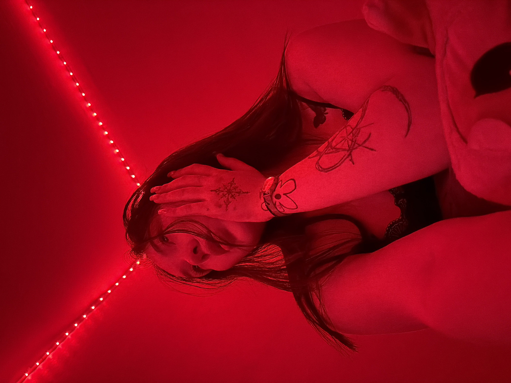 In a dimly lit room, illuminated by a string of red LED lights, a young woman is captured in a low-angle shot. Her head is tilted upwards, with her right hand resting on her forehead and cheek, partially obscuring her face. Her long, dark hair cascades around her shoulders. She appears to be wearing a black lace bralette. Her left arm is visible, displaying tattoos on her forearm, including a star-like design and a floral motif. A watch adorns her wrist. The background is a solid red wall, with the string lights forming an arc above her. The overall impression is intimate and moody, with the red lighting casting a strong hue over the scene. There are no discernible elements that place this image specifically in the city of Chaikovsky, Russia; the location is indeterminable from the visual information provided.