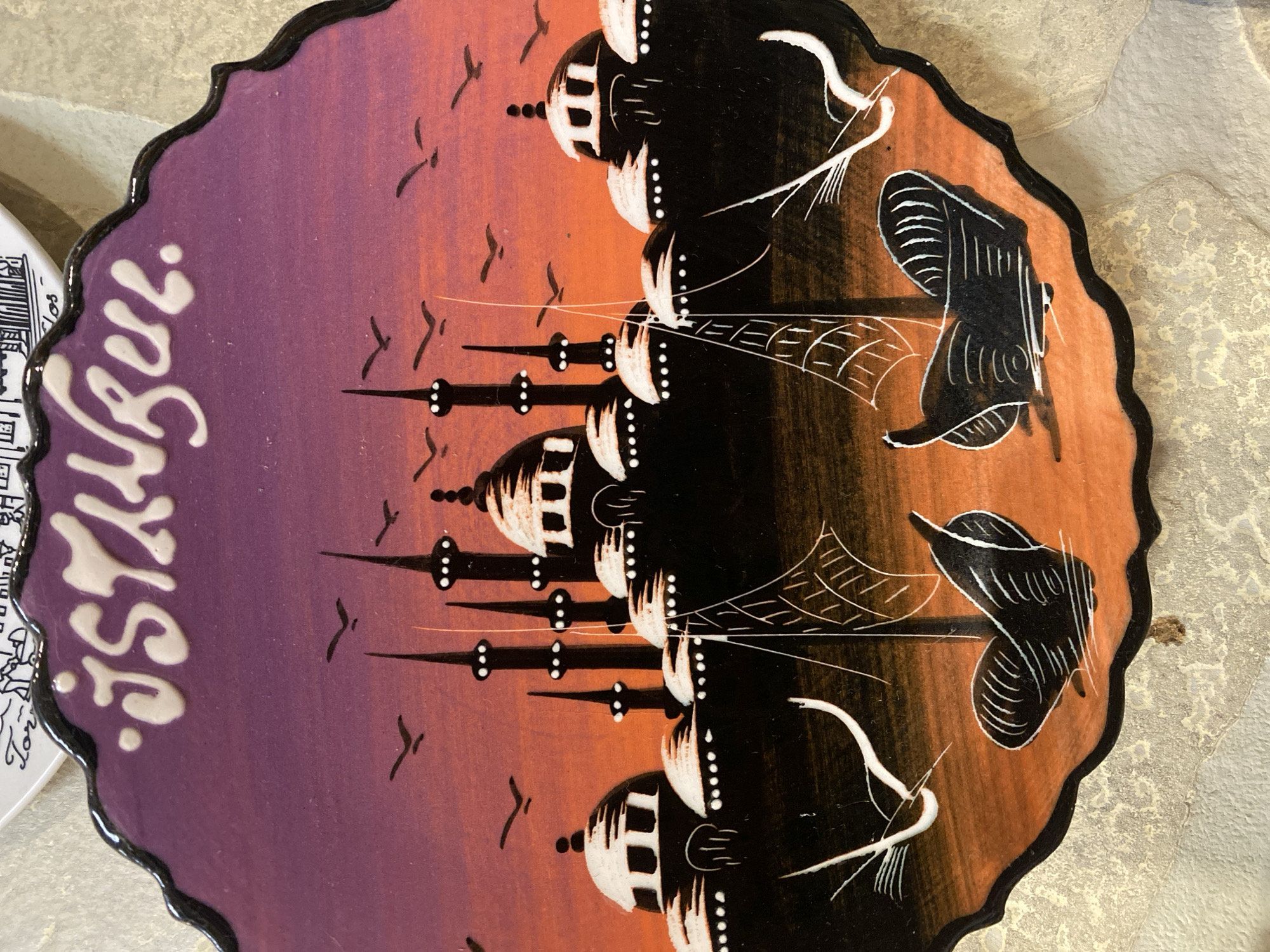 The image shows a decorative ceramic plate with a depiction of Istanbul. The plate is scalloped around the edge with a black border. White lettering at the top spells out "ISTANBUL". The artwork below features silhouettes of mosques and minarets against a gradient sky of purple and orange, transitioning to black at the bottom. Two stylized sailing boats with white sails and dark hulls are depicted in the foreground on orange water. Several small black birds are scattered in the sky. The plate rests on a textured, light-colored surface. The location in the city of Novaya Usman', Russia is not discernible from the image.