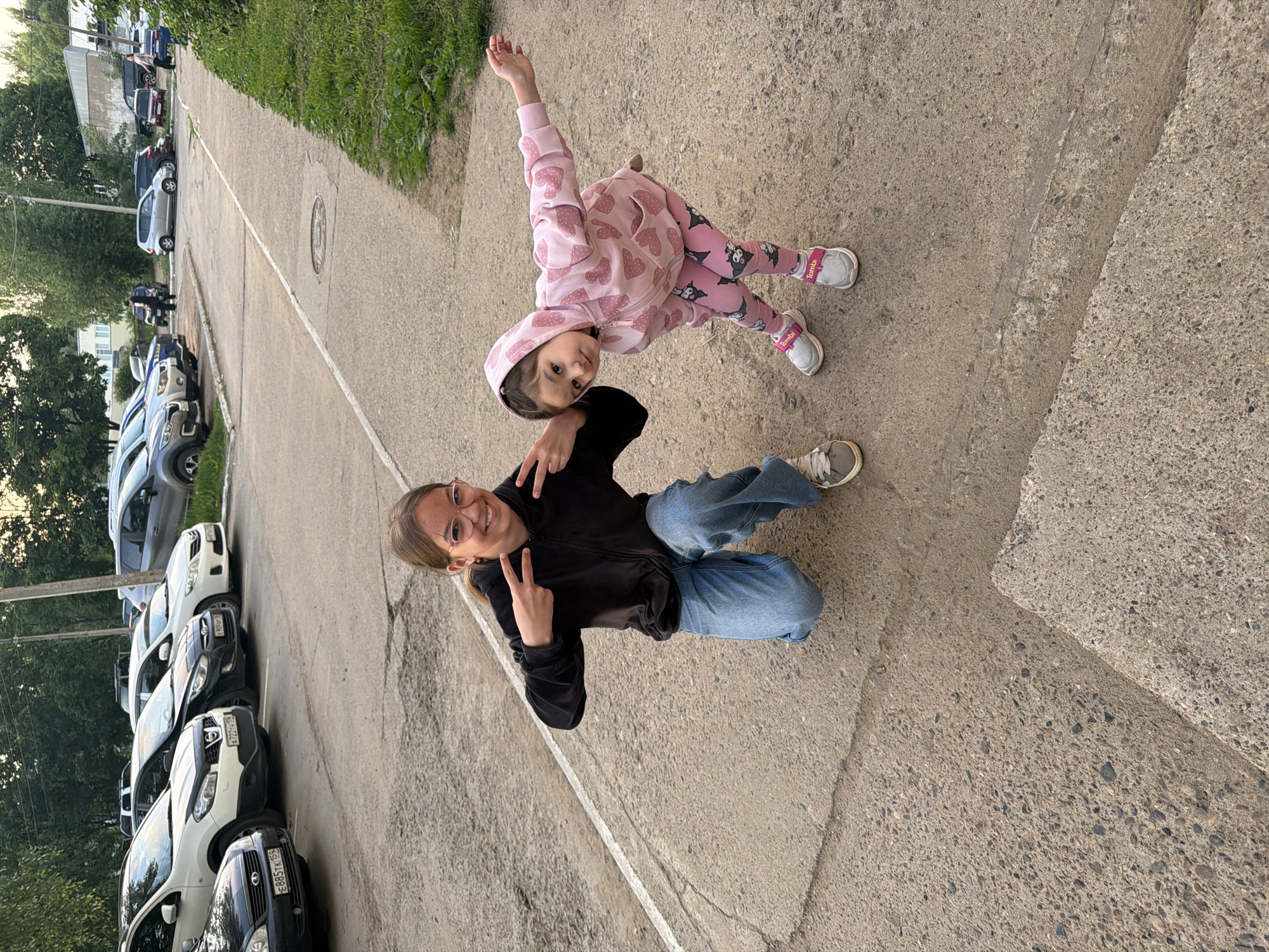 An outdoor, eye-level shot captures a young woman and a child posing on a paved surface. The image is rotated, but the subjects appear to be sitting/standing upright on the ground.
The young woman, with light brown hair and wearing glasses, a black hooded jacket, and blue jeans, is sitting and leaning back slightly, making peace signs with both hands. She is smiling cheerfully at the camera.
Beside her, a young girl, dressed in a pink hooded tracksuit with heart patterns, matching pink and black patterned leggings, and grey sneakers with pink straps, stands with one arm raised, also looking towards the camera with a slight smile.
They are positioned on a cracked asphalt surface with a white painted line. To their left (if the image were rotated correctly), a row of cars is parked along the side of the road. Visible car license plates include "E885TK 152", "A722HH 152", "E010EY 152", and "T160TH 152", which are Russian plates, with "152" indicating the Nizhny Novgorod region. Beyond the cars, trees and distant buildings are visible. To the subjects' right, a strip of grass runs alongside the paved area.
The scene is lit by natural daylight, suggesting late afternoon or early evening. Both individuals appear happy and relaxed. The text "Tombu" is visible on the strap of the child's sneakers.