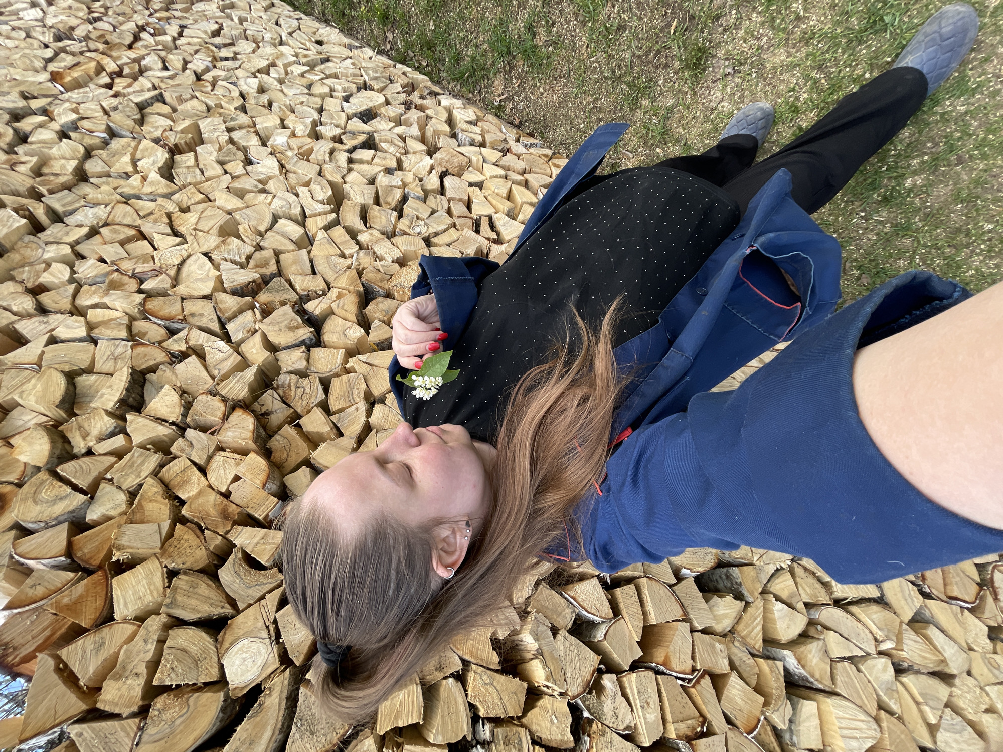 A woman is lying on a pile of split logs. She has brown hair, which is tied back in a ponytail and is wearing a black dress with white polka dots and a blue jacket. She is holding a small cluster of white flowers with a green leaf in her hand. Her eyes are closed. She is also wearing black pants and light blue shoes. The logs are neatly stacked and are surrounded by green grass. The photo appears to be taken from above, possibly as a selfie. The location is in Балдейка, Russia.