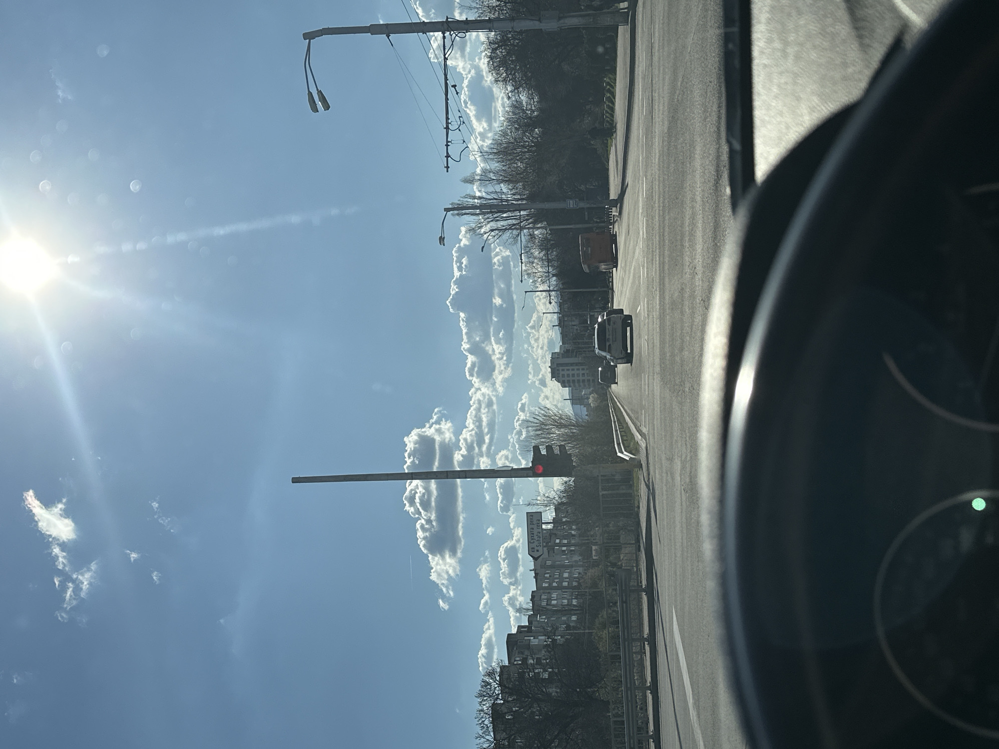 A low-angle, daytime shot taken from the driver's seat of a vehicle shows a street in Sofia, Bulgaria. The view looks forward through the windshield. The sky is blue with numerous cumulus clouds. The sun is bright and causes lens flare. Street lamps and trolley wires are visible on the left. A traffic light is visible in the distance, illuminated with a red light. Various buildings line the street. Two cars and a truck are visible further down the road. The interior of the car, including the steering wheel and dashboard, is partially visible.