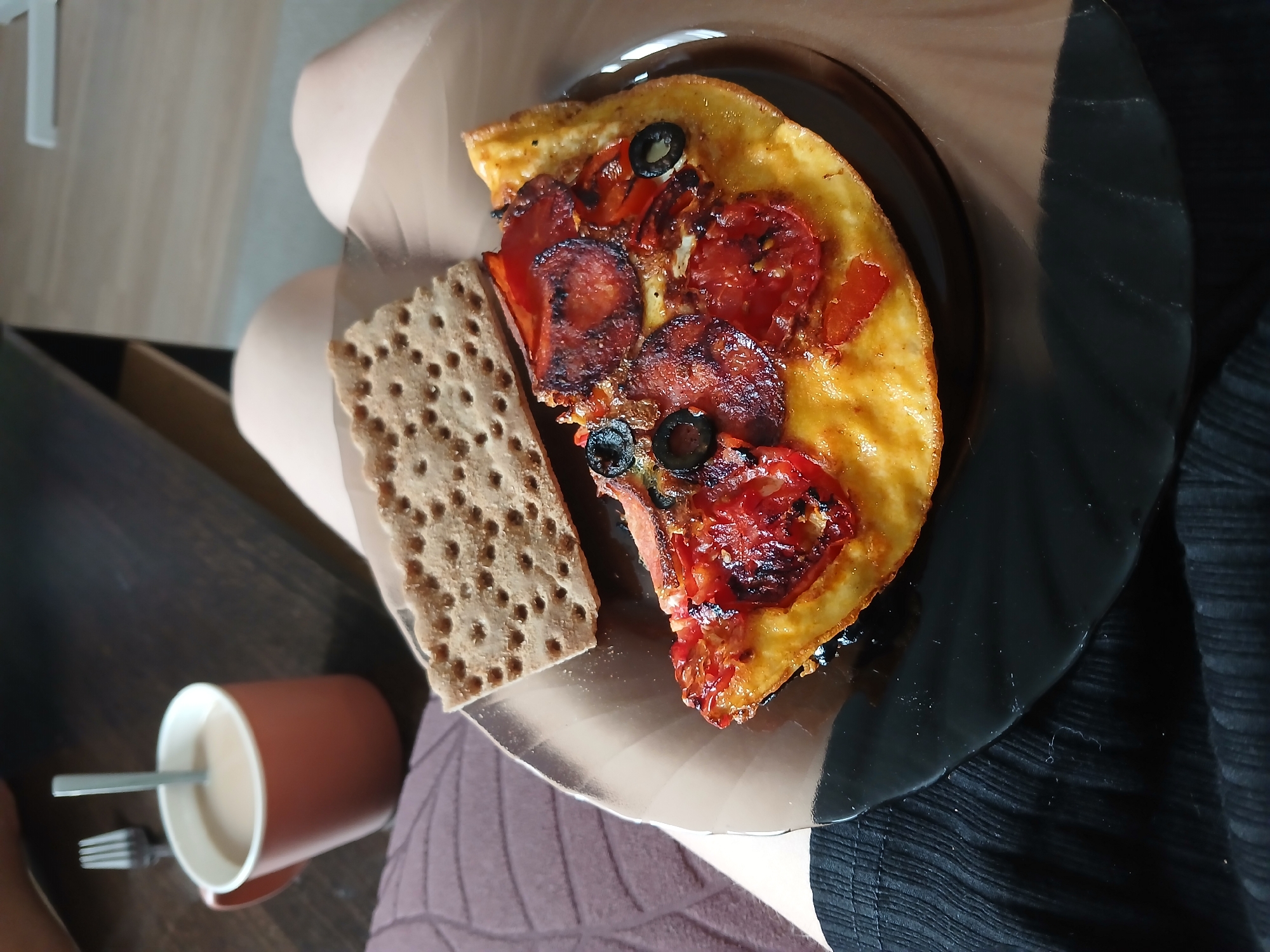 A slice of omelet topped with pepperoni, tomatoes, and olives sits on a dark brown plate, accompanied by a square cracker. A fork and spoon rest beside a mug filled with a light-colored beverage. The scene is set on a dark wooden surface, with portions of someone's legs and dark fabric visible in the foreground. The lighting suggests an indoor environment.