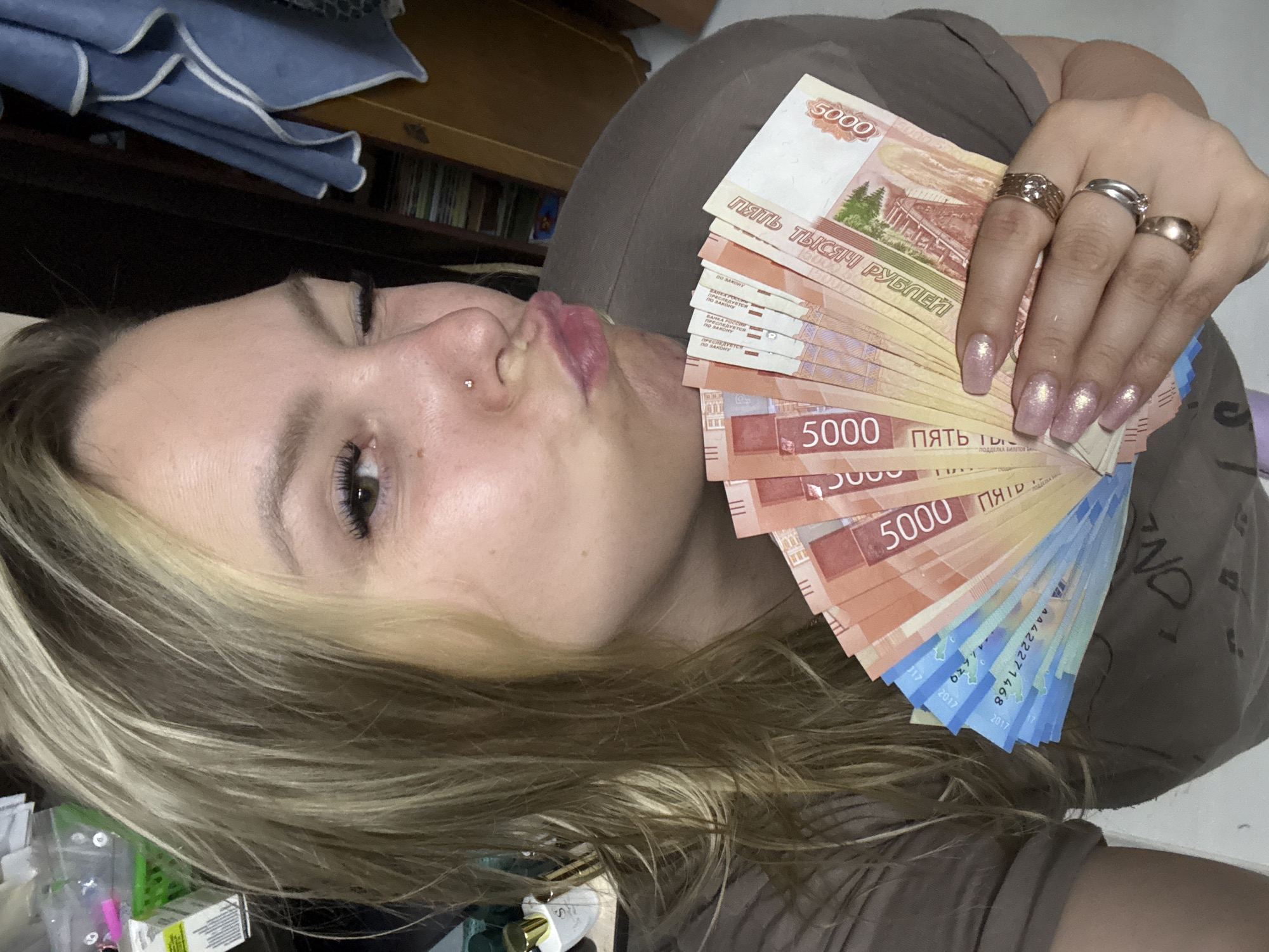 A woman with blonde hair and several rings on her fingers is holding a fan of 5000 Russian Ruble bills. She has a piercing in her nose and is pursing her lips. The photograph appears to have been taken indoors. It is not possible to determine the city of Markovskiy from the image.