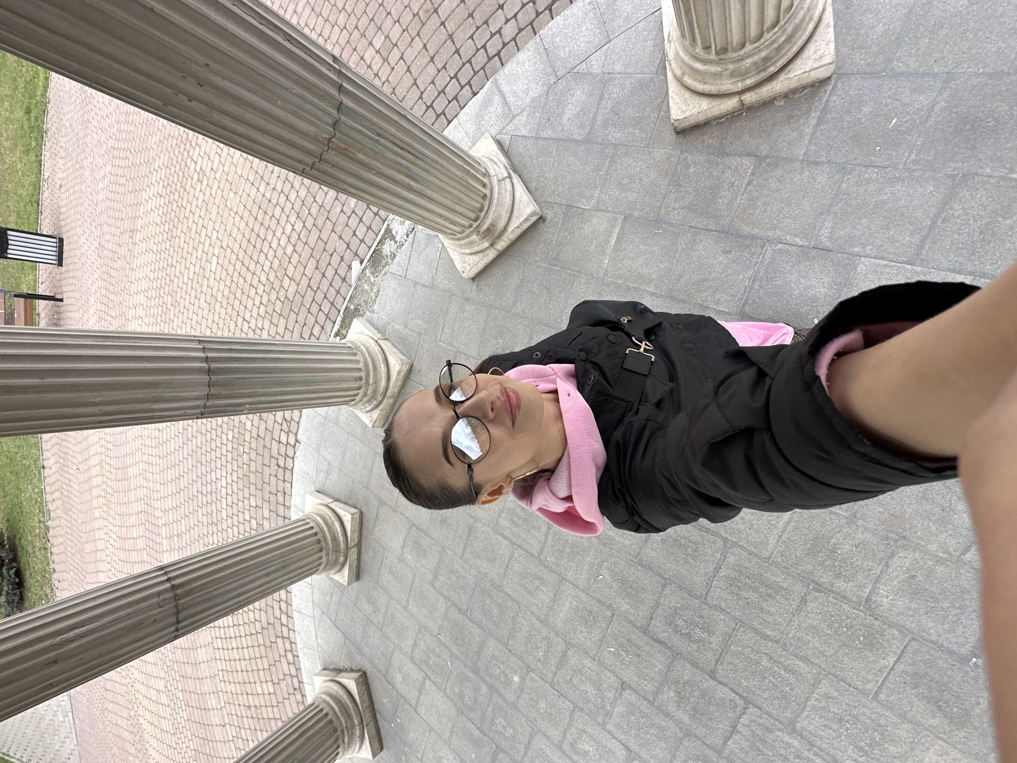 A selfie of a woman with eyeglasses wearing a black jacket and a pink hoodie, standing on a paved surface near a structure with classical columns. The location is likely Yekaterinburg, Russia, given the paving stone and column style common in the city's architecture. A bench and some grass can be seen in the background. The columns surround the woman.