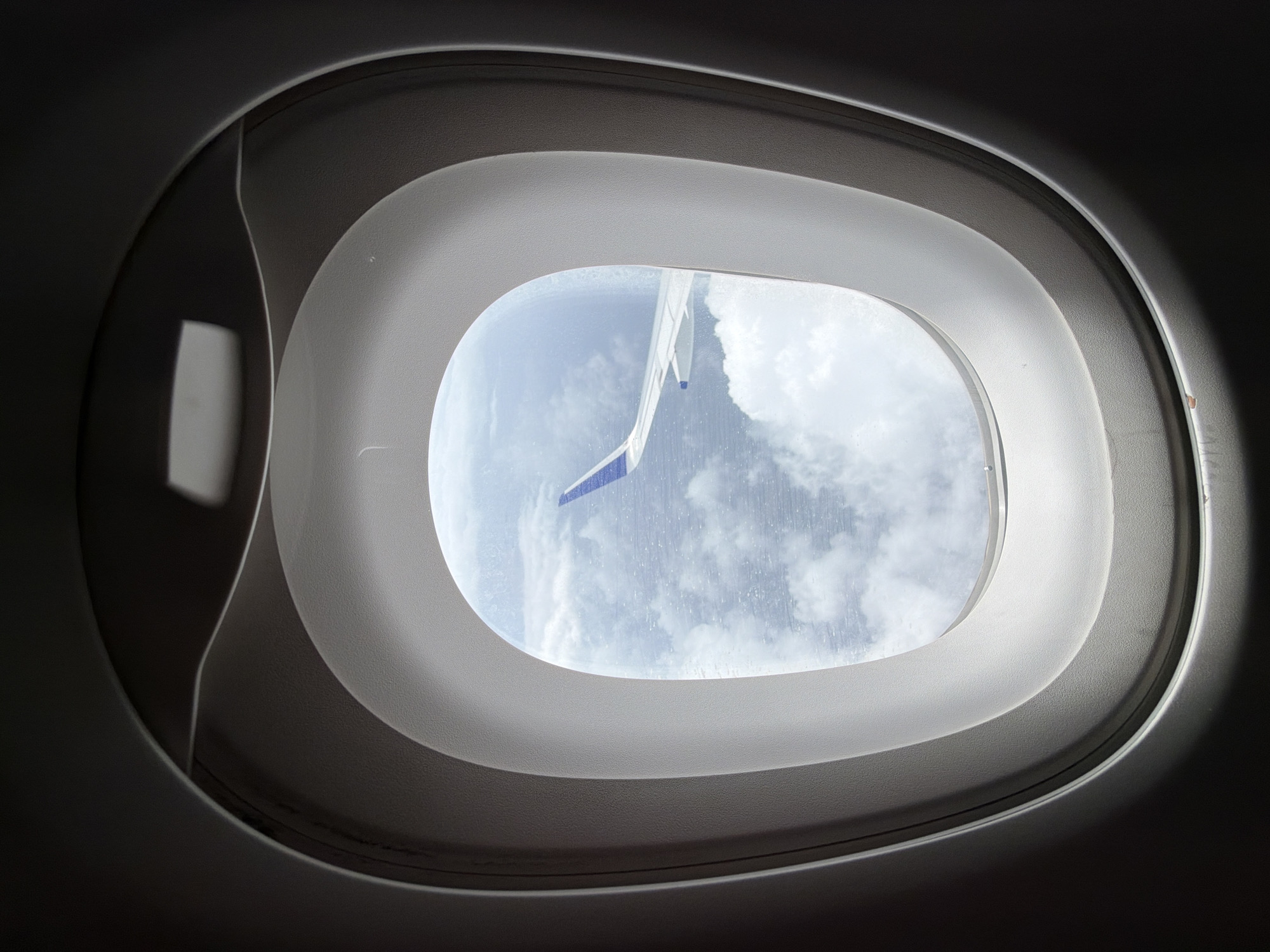 The image shows a view through an airplane window. The window has a distinctive oval shape with layered framing, typical of aircraft cabins. Through the window, we can see a vast expanse of sky filled with white, fluffy clouds. A portion of an airplane wing, with blue and white markings, is visible extending from the right side of the frame into the cloudy sky. The lighting suggests it is daytime, likely mid-flight. There are no people or specific city landmarks visible in this image. Therefore, it is not possible to determine a location within the city of Bhiwandi, India, based solely on this photograph.