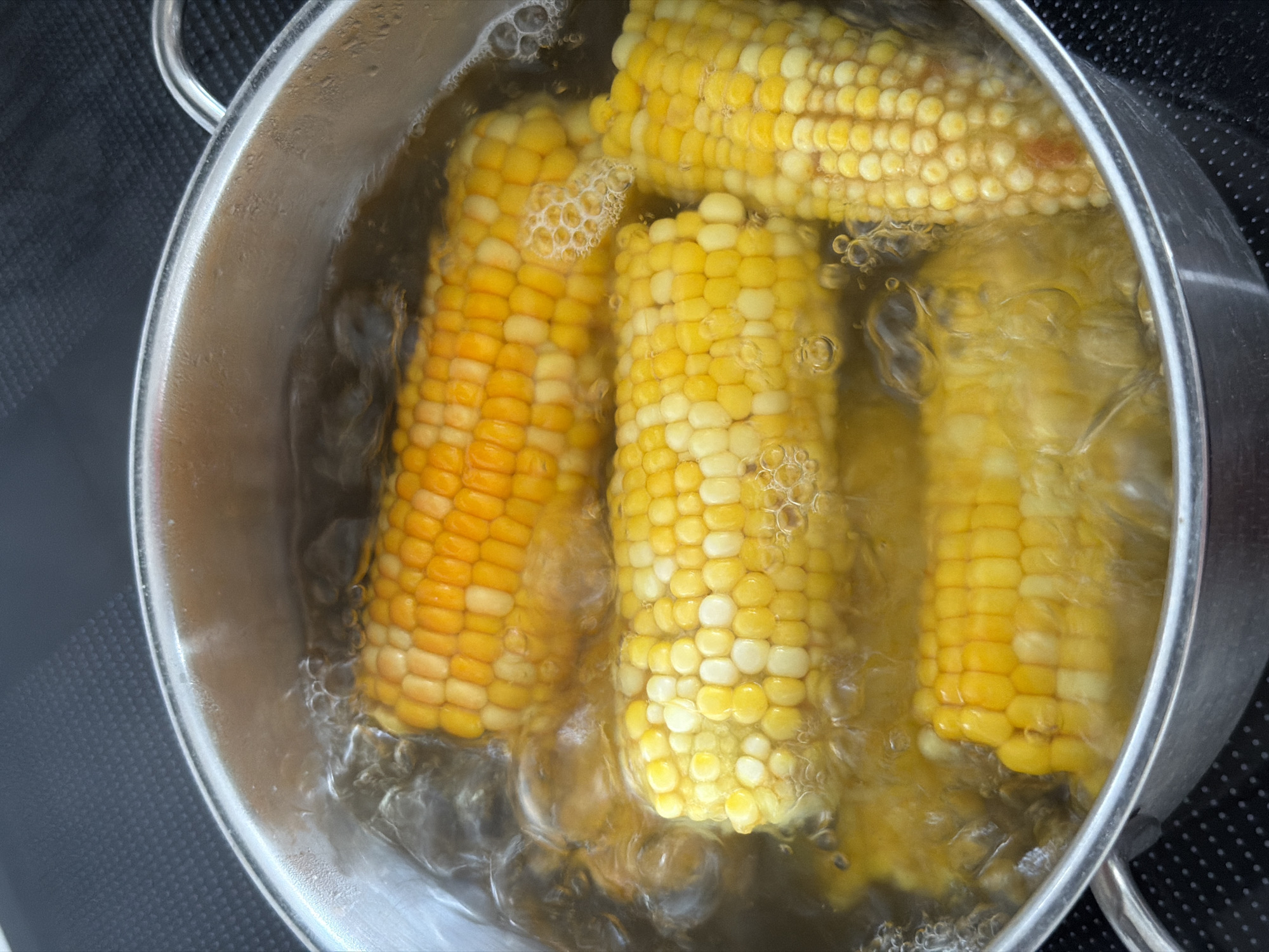 Four ears of corn are boiling in a stainless steel pot filled with water. The corn is a mix of yellow and white kernels. The water is actively boiling, creating bubbles and steam. The pot is on a black stovetop.