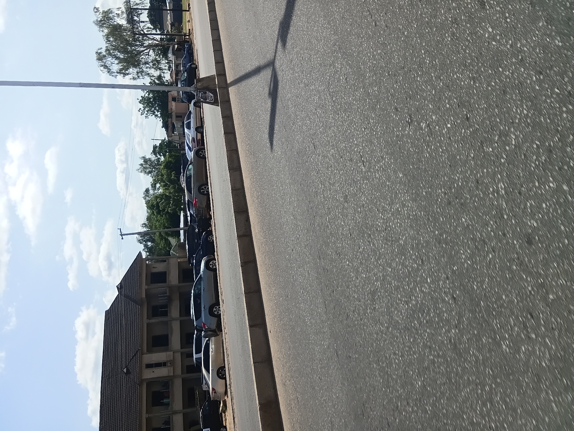 The image shows a street scene in Jalingo, Nigeria, with a building and several cars parked along the side of the road. A light pole is visible in the foreground, casting a shadow on the asphalt. The sky is blue with scattered clouds. The building appears to be a commercial or residential structure with multiple stories and a dark-tiled roof. Cars parked include sedans and SUVs in various colors. The road is paved with asphalt, and a concrete barrier separates it from the sidewalk or parking area. The environment appears to be a typical urban or suburban setting in Nigeria.