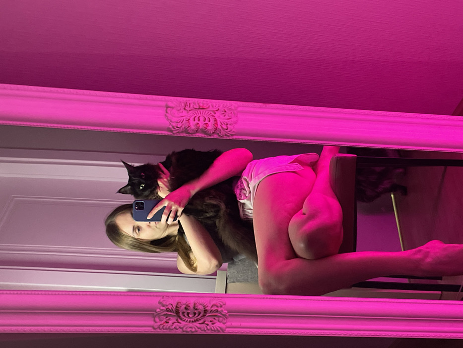 A reflection in a decorative, pink-lit mirror captures a woman holding a dark-haired cat. She holds up a smartphone, presumably taking the picture. Her left arm extends around the cat. Her legs are also visible, with one foot propped up on a chair, creating an angled pose. The image is bathed in pink light, and the mirror's frame is intricately designed.