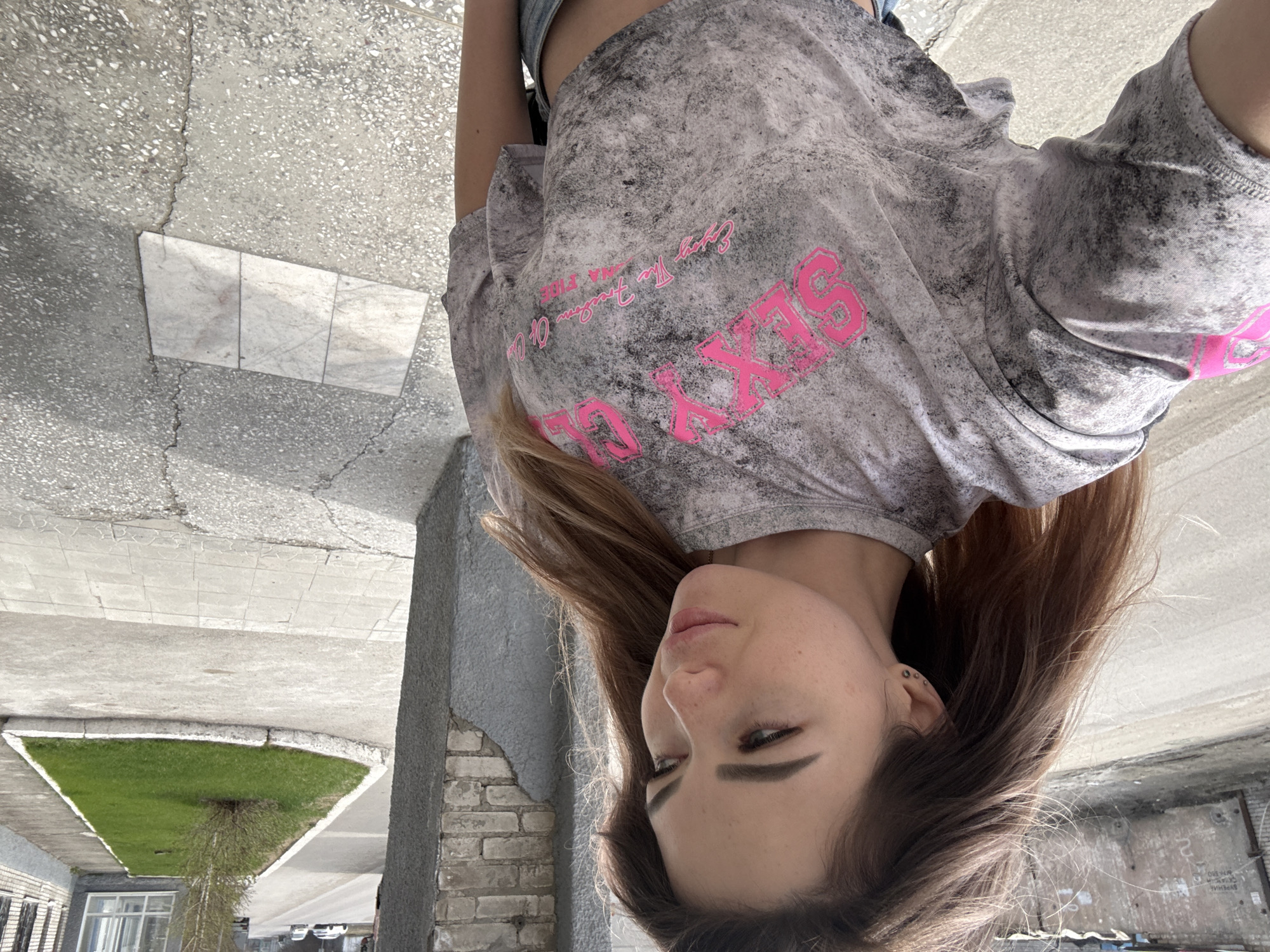 A young woman is facing the camera, with the image rotated upside down. She is wearing a gray t-shirt with the words "Sexy Club" in pink lettering. Her hair is brown and styled. The background appears to be an urban street scene with concrete sidewalks, a small patch of grass, and a building facade. The location is Unknown, Unknown.