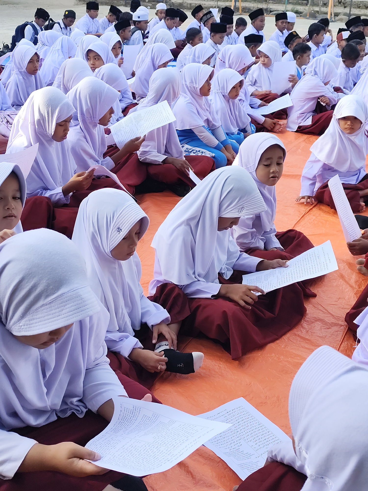 A group of young Muslim students, most of whom are girls wearing white hijabs and uniforms, are sitting on an orange tarp, reading from papers. Some of the boys in the background are also wearing white uniforms and black skullcaps. The students appear to be engaged in a lesson or religious activity. The environment seems to be outdoors, possibly a schoolyard or an open space, with a blurred background of trees and buildings. The lighting suggests it is daytime.