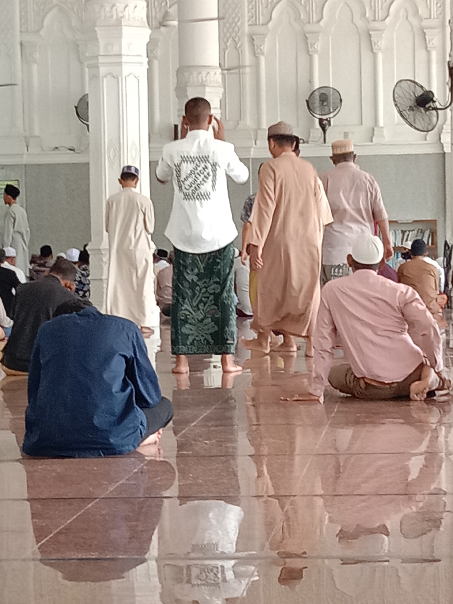 This photograph captures the interior of a mosque, likely in Lhokseumawe, Indonesia, given the attire and setting. The scene depicts several individuals, predominantly men, in various stages of prayer or observation within the mosque. The flooring appears to be polished, reflecting the light and the figures.
In the foreground, a person wearing a dark blue garment is seated on the floor, facing away from the camera. To their right, another individual in a light pink long-sleeved shirt is also seated, with their back turned. Further into the frame, men stand and move about. One man, facing away, wears a white long-sleeved shirt with intricate black text on the back and a green patterned sarong. Another man in a light brown robe walks away from the camera, accompanied by others in similar attire. The architectural elements of the mosque are visible, including white pillars with decorative carvings and arched alcoves. Two industrial-style fans are mounted on the wall in the background, suggesting a warm climate. The lighting is soft and diffused, indicating natural light filtered through the mosque's openings. The overall atmosphere is one of quiet reverence and communal gathering.