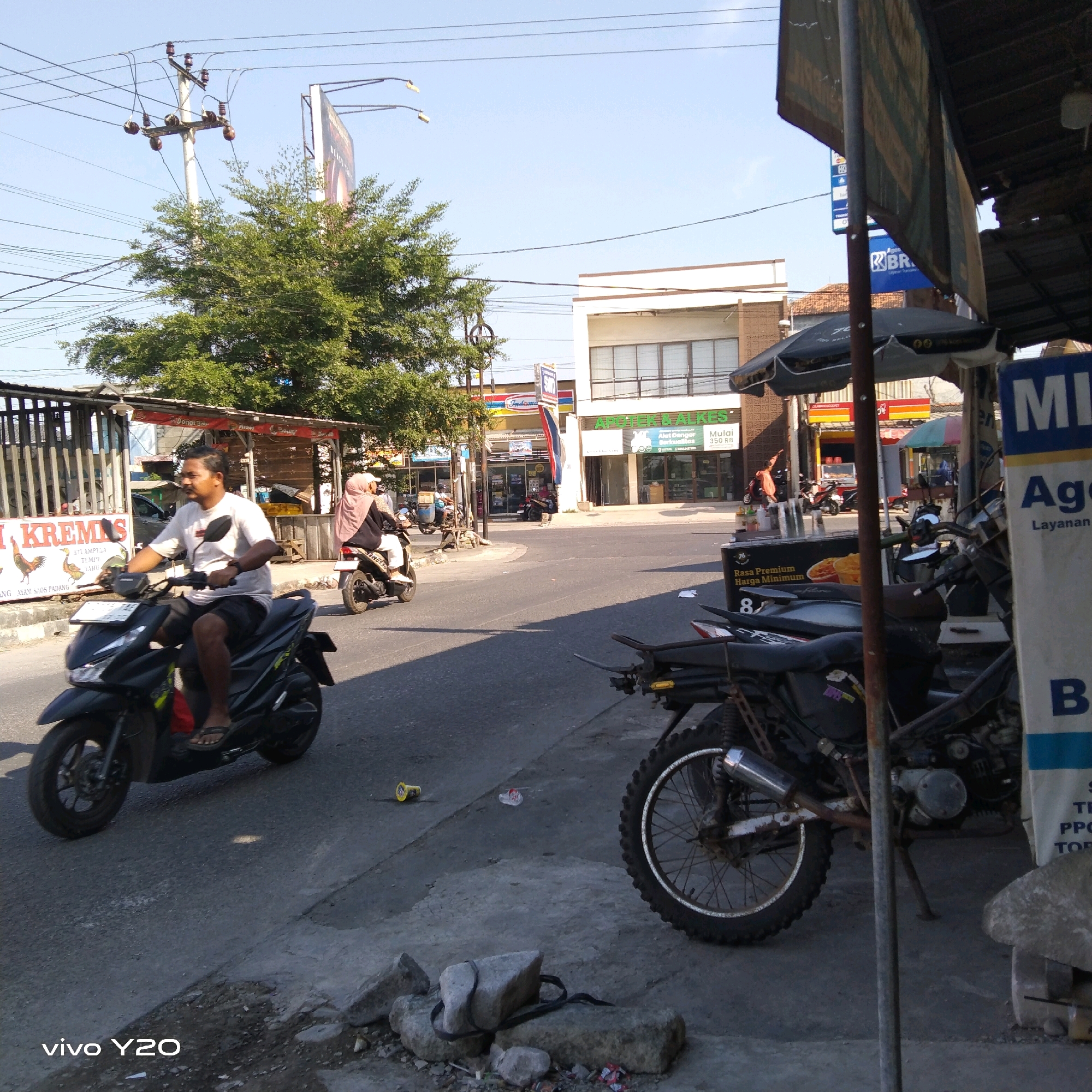 The image depicts a street scene in an urban environment. In the foreground, a black motorcycle is parked, with a portion of another motorcycle visible to its right. The street is paved, with some debris scattered on the side. Further back, a man on a black scooter is riding down the street, wearing a white t-shirt and shorts. Behind him, another scooter carries a person wearing a hijab. On the left side of the street, a building with a fence and a sign that reads "KREMIS" is visible. Across the street, a commercial building with multiple windows and a sign that says "APOTIK ALKES" can be seen. There are also power lines and a tree in the background. The sky is clear and blue. The overall impression is that of a typical day in a bustling city.