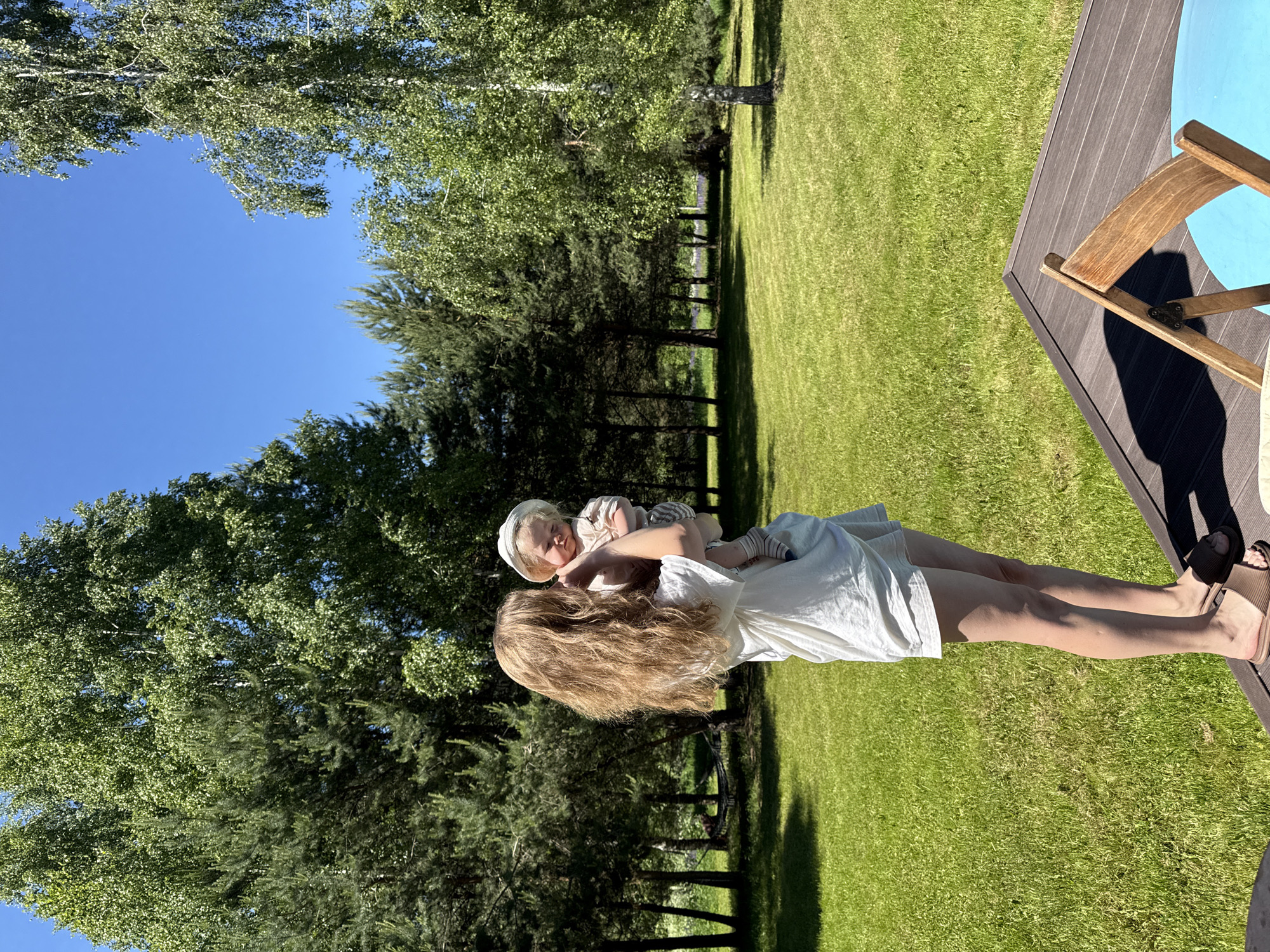 A young adult female is depicted standing outdoors holding an infant under a clear blue sky. The female, positioned on a dark brown slatted deck in the foreground, faces slightly away from the camera, looking towards the infant. She has long, wavy, light brown hair and is dressed in a loose white top, white shorts, and dark brown slide sandals. The infant, wearing a light-colored sun hat, a light top, striped shorts, and striped socks, is held facing forward, looking slightly downwards with eyes partially closed. The background consists of a sprawling, neatly mowed, vibrant green lawn and a dense arrangement of trees, including tall dark green conifers and lighter green birches with white bark. Strong sunlight from the left casts distinct shadows on the grass and the deck. A portion of a wooden folding chair with a light blue cushion is visible on the deck in the lower right. In the mid-ground, a dark-colored hammock is discernible, strung between two trees amidst the shaded area. The scene is located in the city of Копнино, Russia.