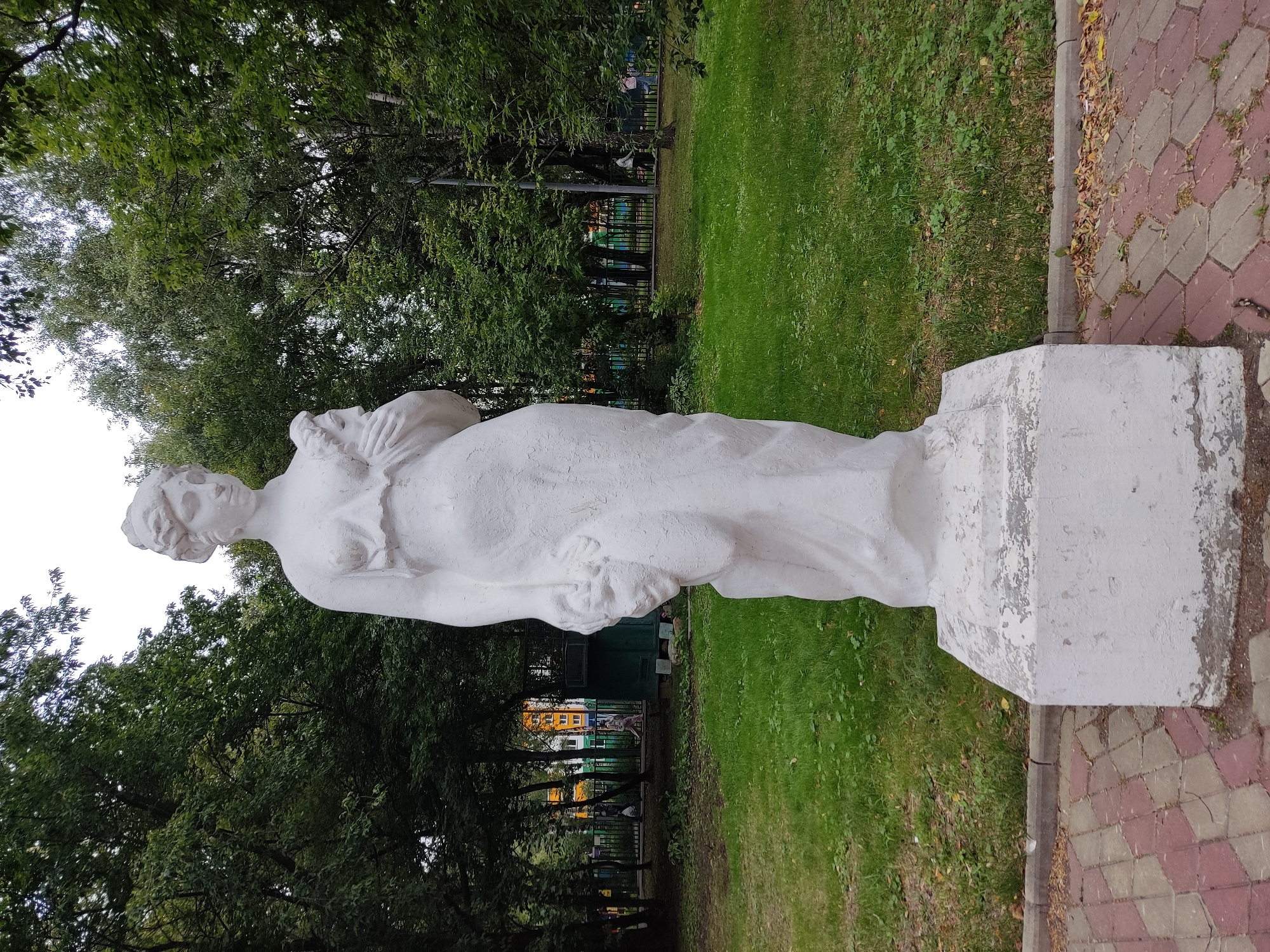 A white, classical-style statue of a woman stands in a park. The statue is of a woman wearing a draped dress, with her hair styled up. She is holding a bundle of what appears to be flowers or fruit in her left arm and a second bundle in her right hand, which is lowered. The statue is positioned on a rectangular, multi-layered pedestal that shows signs of wear and peeling paint.
The statue is situated on a paved walkway composed of red and gray hexagonal tiles. To the left of the statue, a lawn area with sparse green grass and scattered fallen leaves is visible. In the background, a dark green metal fence encloses a playground area with colorful structures and a building with an orange facade. Mature trees with dense green foliage surround the park, creating a shaded environment. The sky above is overcast and pale. The scene is located in the courtyard of residential buildings at the address 15 Komsomolsky Prospekt, Moscow, Russia.