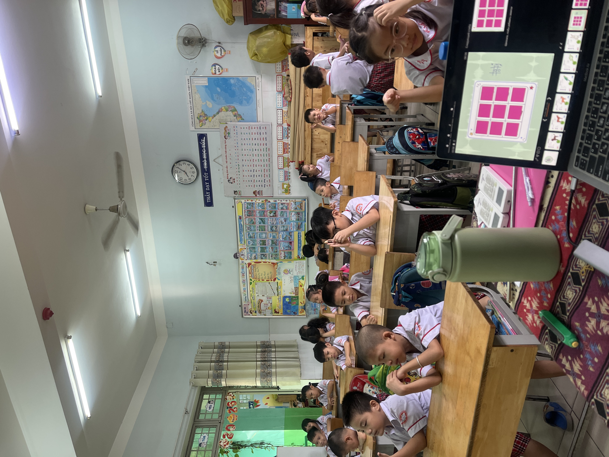 The image shows a classroom in Thuận An, Vietnam, filled with elementary school students in white and red uniforms sitting at wooden desks. Many are resting their heads on the desks. Educational posters and maps decorate the walls, along with a clock displaying 10:13. A laptop displaying a game is visible in the foreground. The room is well-lit by fluorescent lights, and a ceiling fan hangs in the upper left corner.