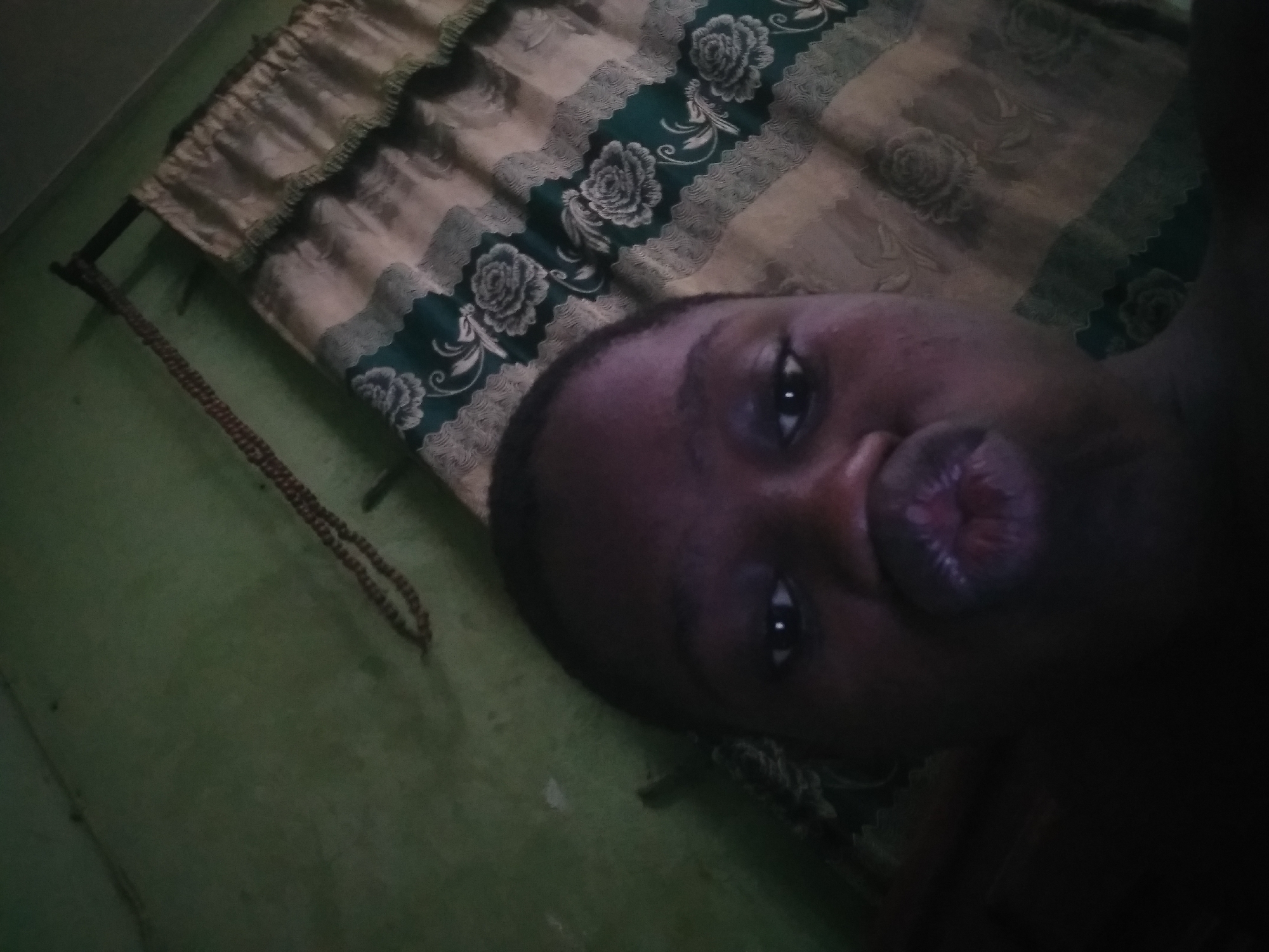 The image shows a dark-skinned person lying on a surface with a green wall in the background. A curtain with floral patterns is visible above the person. A string of beads hangs to the left. The photograph appears to be taken indoors. Location: Shangisha, Nigeria.