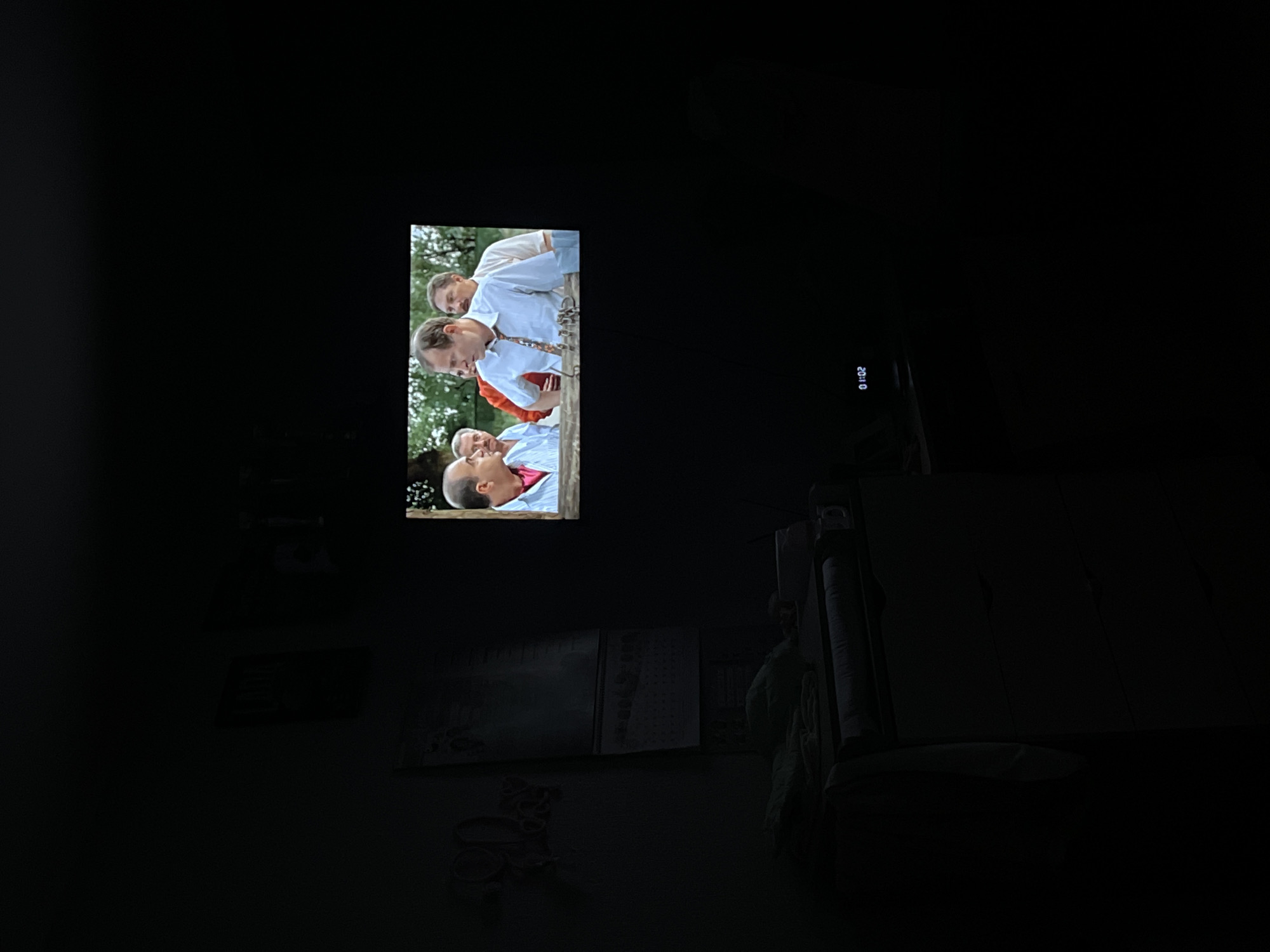 The image is a dimly lit indoor shot of a room with a television displaying a scene of several men in shirts and ties. The TV screen shows four men outside, seemingly engaged in a conversation or discussion. The timestamp "20:10" is visible in the corner of the screen. In the foreground and surrounding areas of the room, details are difficult to discern due to the low light conditions. A couch and other furniture are vaguely outlined in the darkness. Without any additional evidence, it is impossible to connect the image with the location in the city of Chaikovsky, Russia.