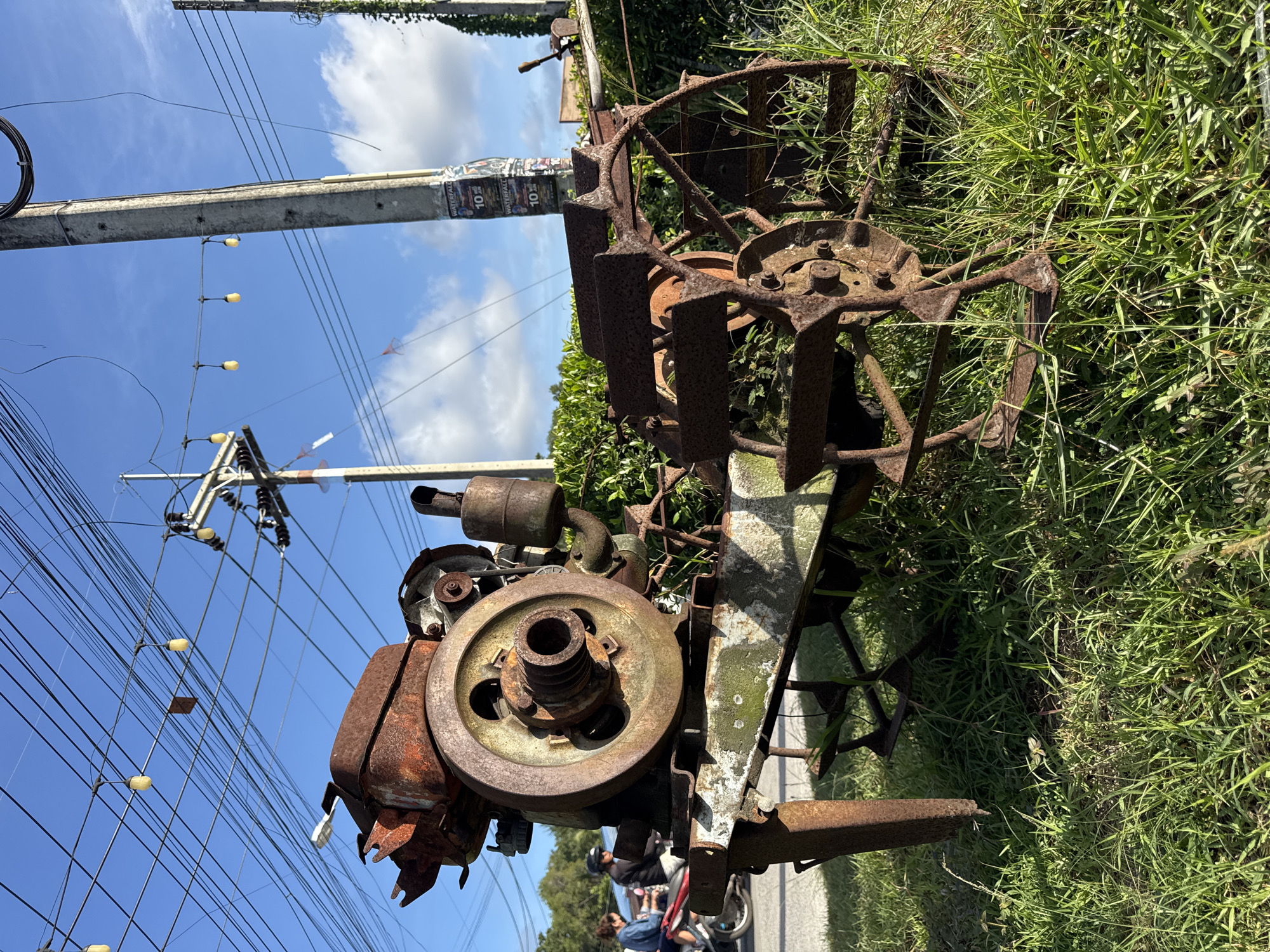 An old, rusty tillers is displayed in front of a lush green bush and tall grass. The tiller has a visible engine and wheels. In the background, there are power lines, utility poles, and a few people on motorcycles passing by on a street. The sky is bright blue with scattered clouds. The location is ตำบลเกาะพะงัน, Thailand.