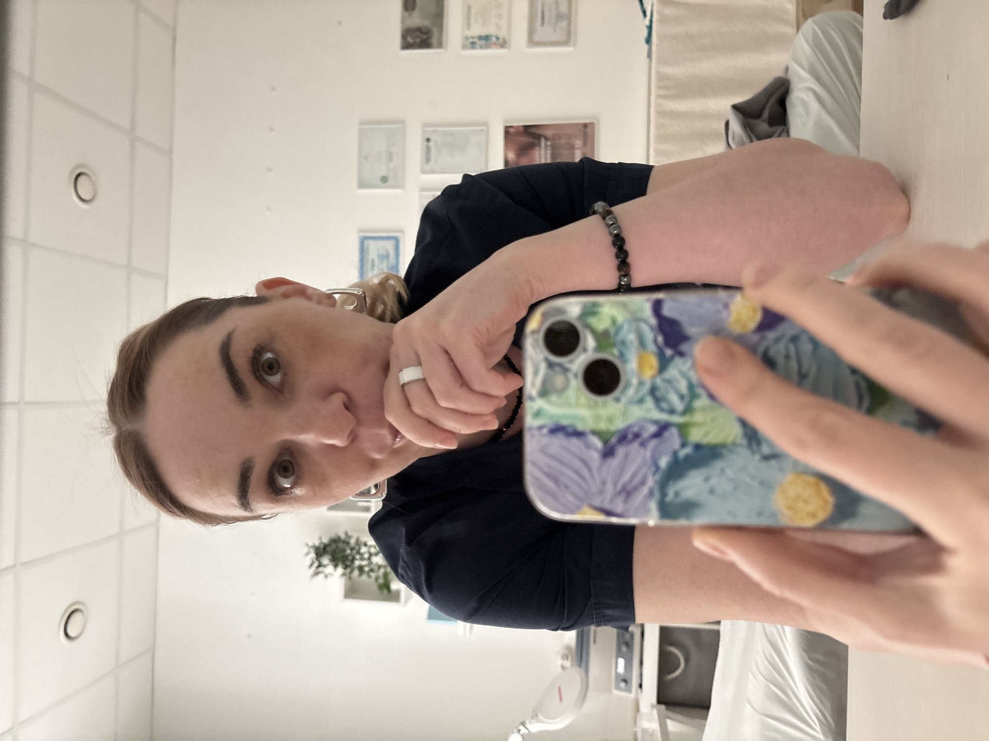 A woman is taking a selfie in a room with white walls and ceilings. The room appears to be a medical office, based on the equipment and certificates visible in the background. The woman is wearing a dark blue top and has light brown hair pulled back. She has a bracelet on her left wrist and a ring on her left hand. She is holding her phone, which has a floral pattern case, to take the picture. The exact location in Vsevolozhsk, Russia cannot be determined from the image alone.