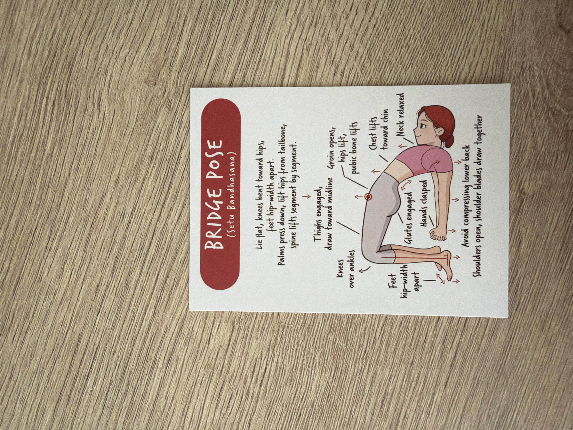 The image shows a yoga pose card featuring the "Bridge Pose" (Setu Bandhasana). The card has a red header with white text and is placed on a wooden surface with a visible wood grain pattern. The illustration depicts a woman in the bridge pose, with arrows and text indicating key alignment points such as knees bent toward hips, feet hip-width apart, palms pressing down, lifted hips, engaged glutes, and shoulders open.
The image is a close-up shot of a card placed on a wooden desk. The background consists of the texture of the wooden surface. There are no elements in the image that would allow for the determination of its location in Pazardzhik, Bulgaria.