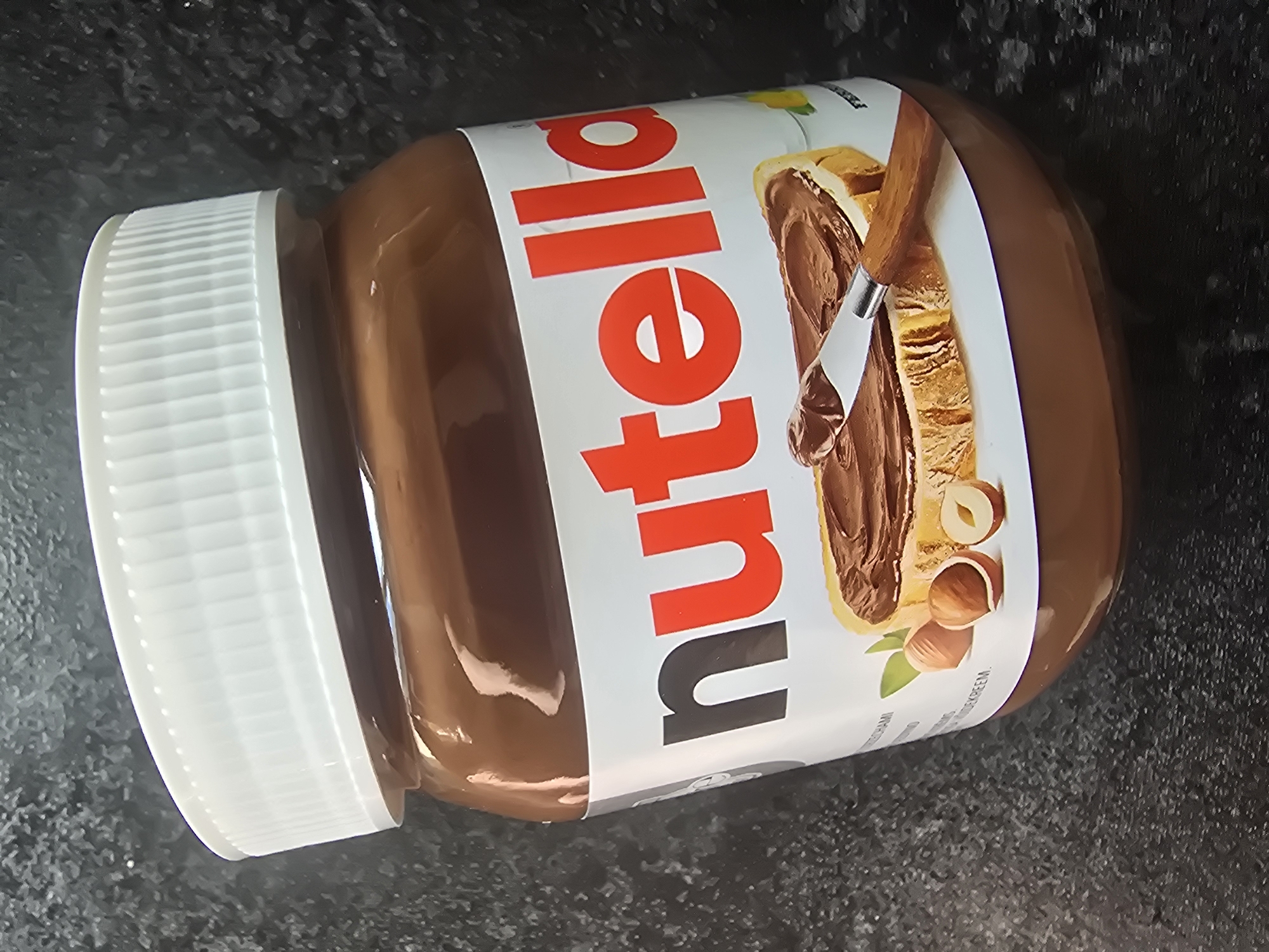 A jar of Nutella sits on a dark, textured surface. The jar has a white lid and a label that prominently features the "Nutella" logo in red and black lettering. Below the logo, an image depicts a slice of bread spread with Nutella, with a knife resting on it. Several hazelnuts are also visible on the label. The background is dark and appears to be a countertop or similar surface with a speckled texture. The lighting is somewhat dim, with reflections visible on the shiny surface of the Nutella jar.