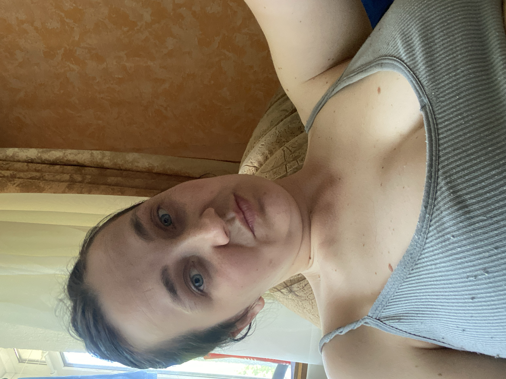 A close-up, vertically oriented selfie shows a person's face and upper body. The person appears to be looking directly at the camera with a neutral expression. They have light-colored skin, blue eyes, and dark brown hair. They are wearing a gray, ribbed tank top. The background consists of a textured orange wall and a glimpse of a window with white curtains. The lighting suggests it is daytime, likely indoors. The image is slightly blurry, particularly around the edges. No text is visible in the image.