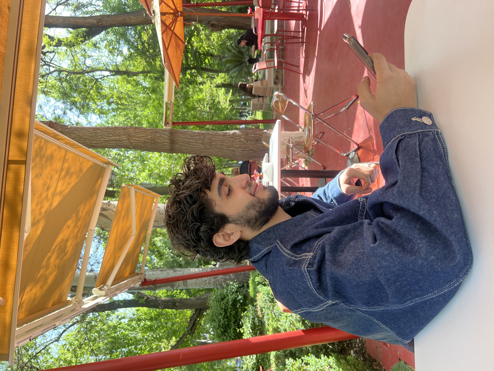 A man is sitting at an outdoor cafe, holding a mobile phone. He has dark curly hair and a beard, wearing a blue denim jacket. The cafe is outdoors, with red-painted surfaces, tables, and chairs. There are umbrellas providing shade. Trees surround the cafe, and other people are in the background. The location is in a city of Unknown, Unknown.