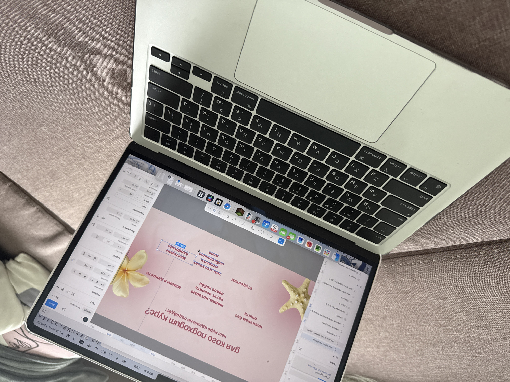 The image shows a close-up of an open silver Apple MacBook laptop resting on a textured, rose-colored fabric surface, likely a couch. The laptop screen displays a design software interface, possibly Figma, with a pink background and Russian text elements. Text boxes on the screen contain phrases like "для кого подходит курс?" (who is the course for?), "наш курс идеально подойдет:" (our course is ideally suited for:), and lists of target audiences. A starfish and a yellow flower are superimposed on the design as visual elements.
On the left side of the screen, a layers panel is visible, showing various design elements. The keyboard of the laptop is clearly visible, with Cyrillic and Latin characters on the keys. The trackpad is also visible. The overall lighting suggests an indoor setting, possibly a home environment.
While the image is a close-up of a laptop, there are no indications or elements within the image that place it specifically in the city of Коробицыно, Russia. The description is purely based on the visual content presented.