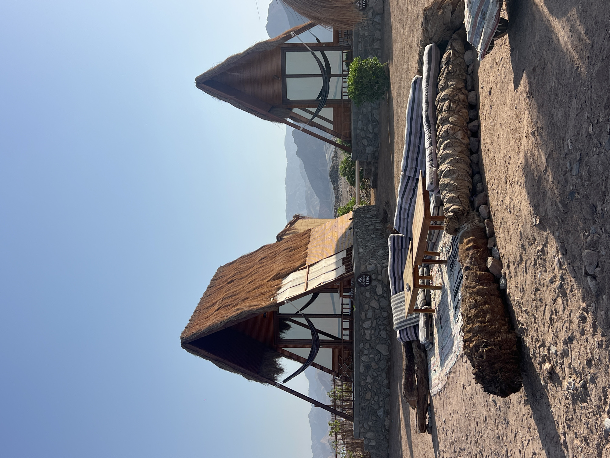The image depicts an outdoor seating area and two A-frame cabins in a desert landscape, likely in Nueiba, Egypt. The cabins have thatched roofs and wooden exteriors with large glass windows, suggesting a glamping or eco-resort setting. Hammocks are visible inside the cabins. In the foreground, there is a low wooden table surrounded by floor cushions and a striped sofa. A large, rough-textured object, possibly a bundle of dried vegetation or wood, lies in front of the seating area. The ground is sandy and rocky, with scattered stones. In the background, mountains are visible under a clear blue sky. The scene suggests a tranquil, rustic environment. There are no people visible in the image.