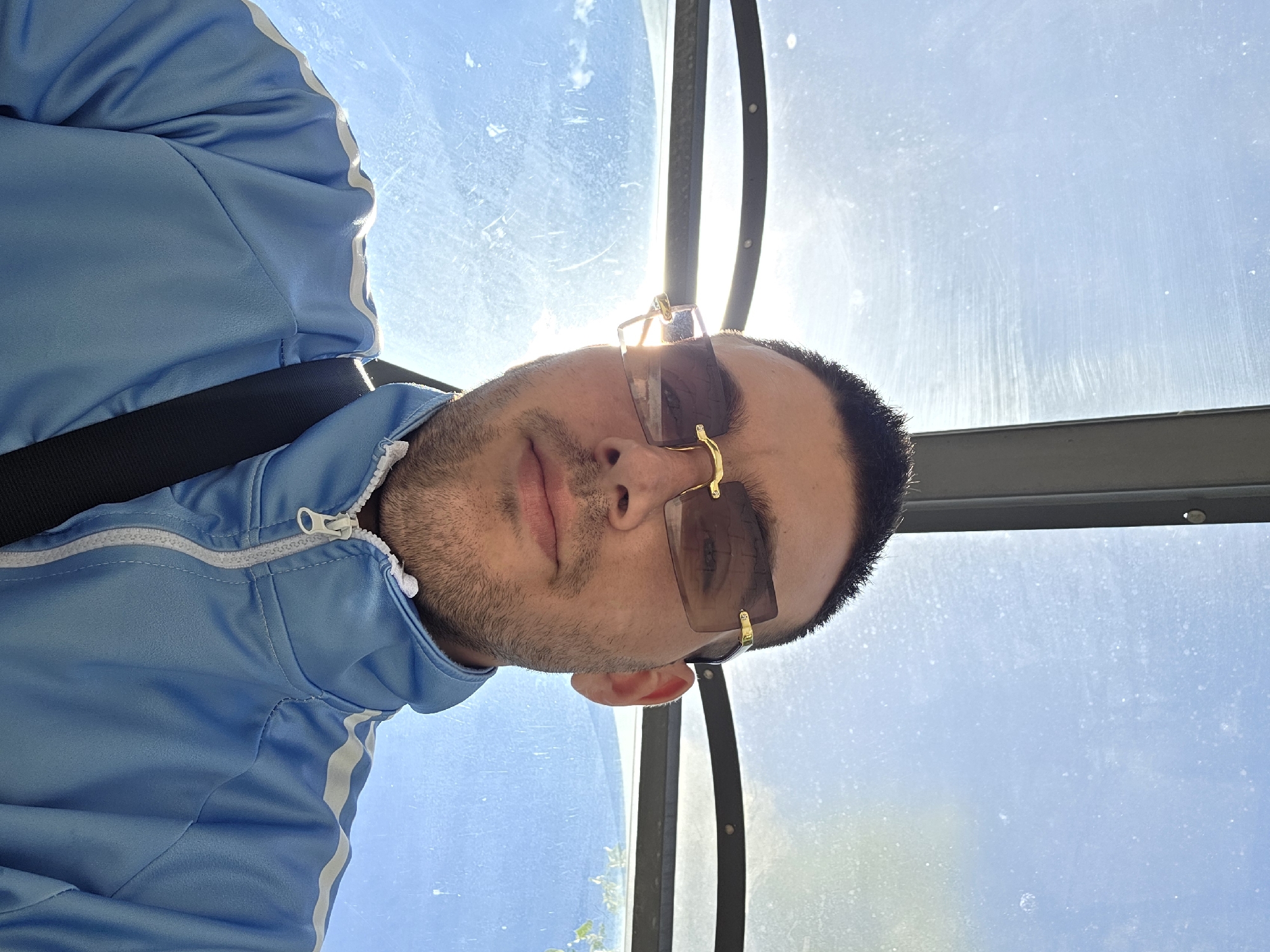 A man with short, dark hair and stubble is looking at the camera. He is wearing rimless, gold-accented sunglasses and a light blue track jacket with white stripes on the sleeves. The jacket is zipped up to the neck. A black strap is visible across his chest. The background is a bright, hazy blue sky with a sun flare. The image appears to be taken outdoors, possibly in a covered area such as a bus stop or shelter, due to the presence of a gray metal structure overhead.