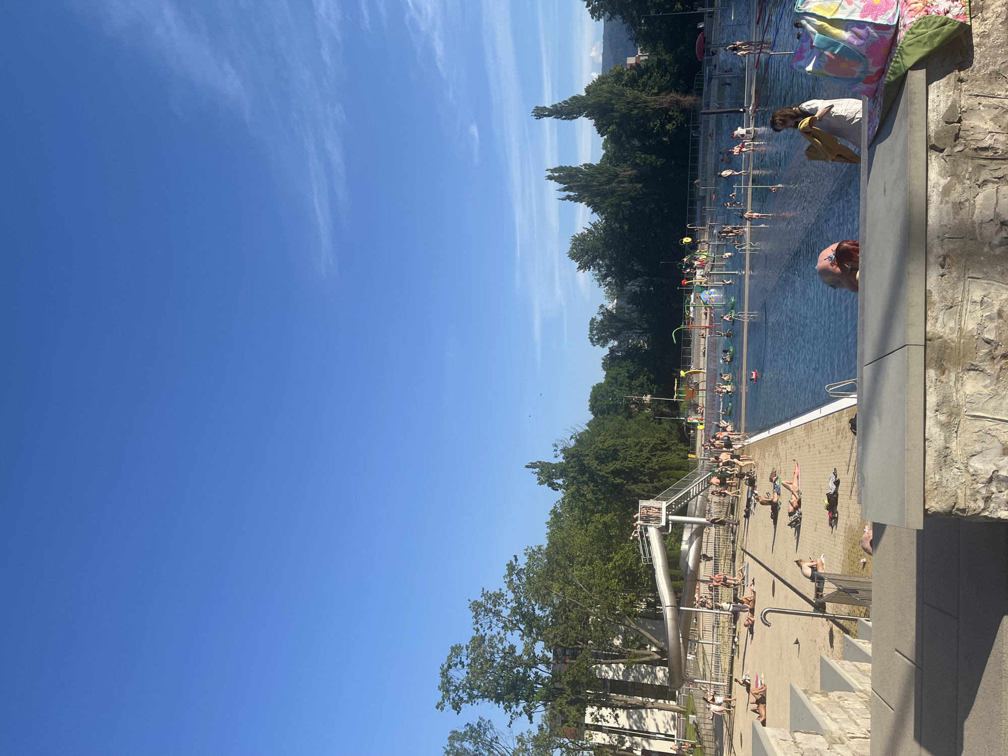 An outdoor swimming pool complex is depicted on a clear, sunny day with a bright blue sky. Numerous people are present, engaging in various activities. Some are sunbathing on the paved area surrounding the pool, while others are in the water, swimming or playing. A large waterslide is visible, with people ascending the stairs and preparing to slide down. In the background, lush green trees line the perimeter of the complex, with buildings and hills visible beyond. The scene suggests a lively atmosphere at a public recreational facility.
