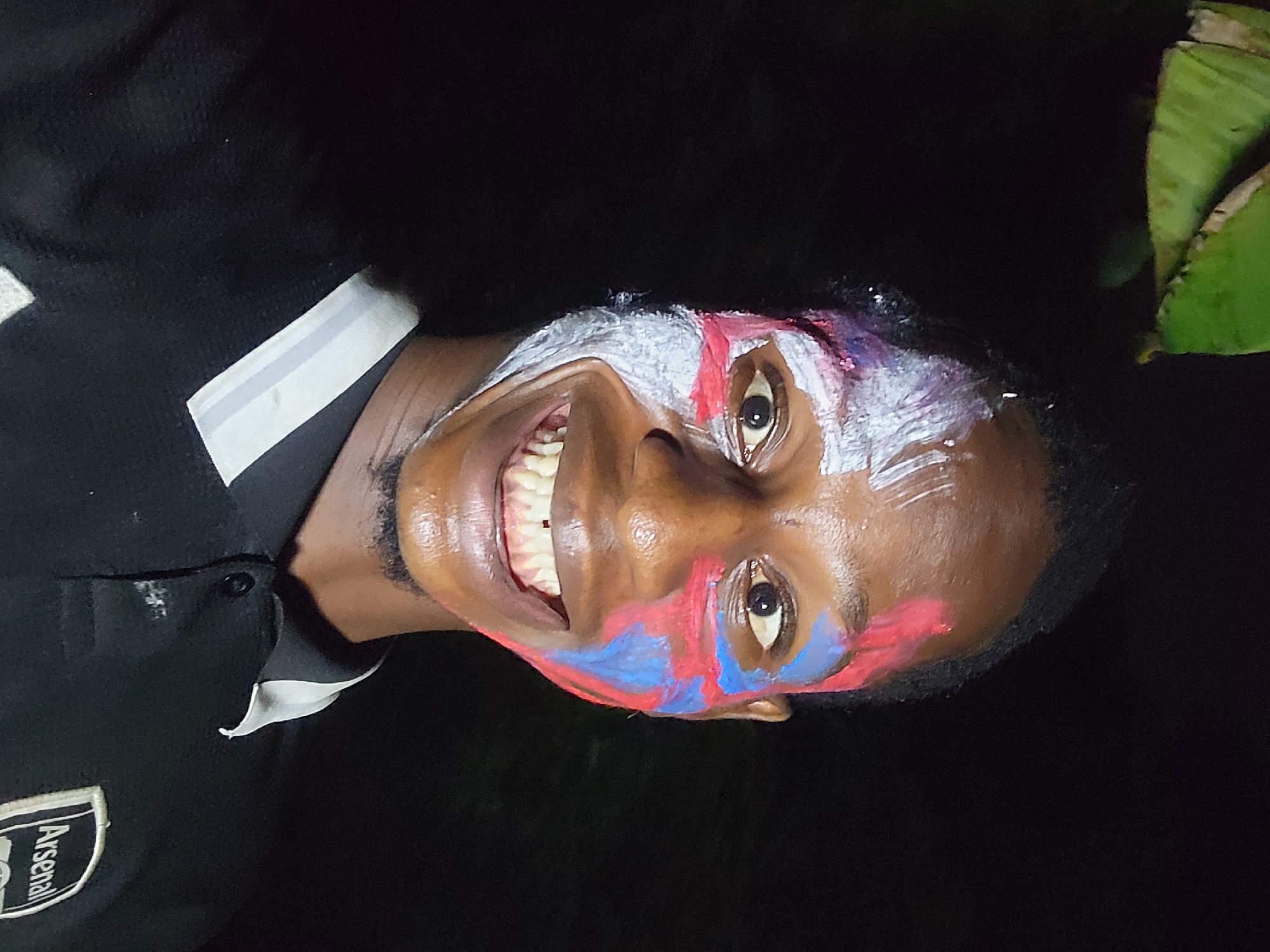 An eye-level, medium shot of a man wearing face paint and an Arsenal polo shirt. The man's face is painted with white streaks across his forehead and red and blue paint around his eyes and cheeks. He is smiling broadly, revealing his teeth. The background appears dark, with green foliage visible in the top right corner. The location is Nkpor, Nigeria.