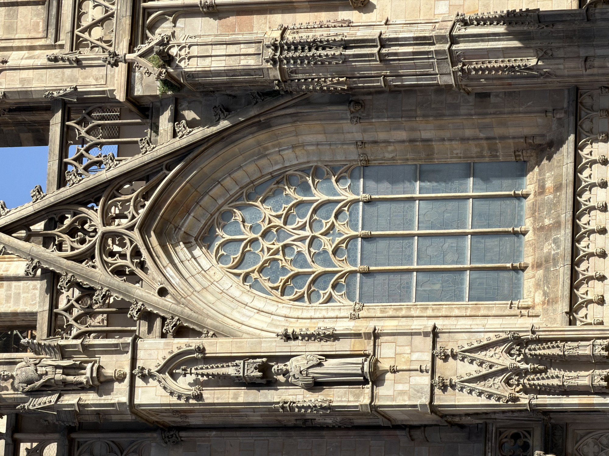 The image shows a close-up view of a Gothic facade of a large stone building, likely a cathedral, in Barcelona, Spain. The architecture features intricate stone carvings, pointed arches, and a large stained-glass window with a tracery design. Statues of figures in religious attire are integrated into the facade, including one of a monk holding a book, positioned in a niche to the left of the central window. The stonework is weathered, with some areas showing signs of age and lichen growth. The sky is visible at the top of the image, a clear blue. The lighting suggests it is daytime, with strong sunlight casting shadows and highlighting the three-dimensional details of the architecture. The overall impression is one of historical grandeur and religious significance.