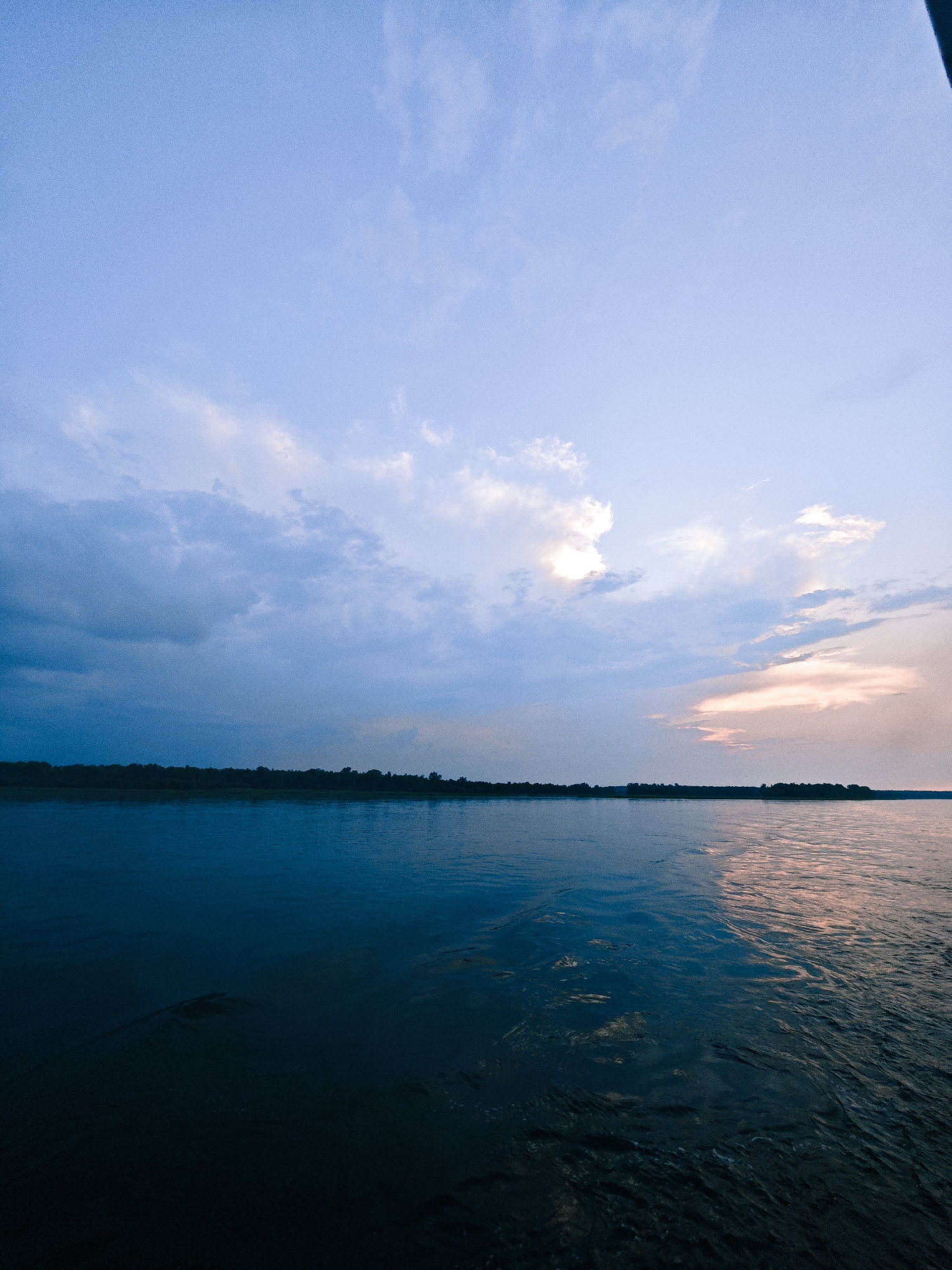 The image depicts a wide, calm river at dusk. The sky is a gradient of blues and purples, with scattered clouds tinged with pink from the setting sun. A dark, tree-lined shoreline stretches across the horizon. The water is dark and reflective, with subtle ripples indicating movement. The scene appears serene and natural. This is a scene of the Angara River in Kazachinskoye, Russia, at sunset.