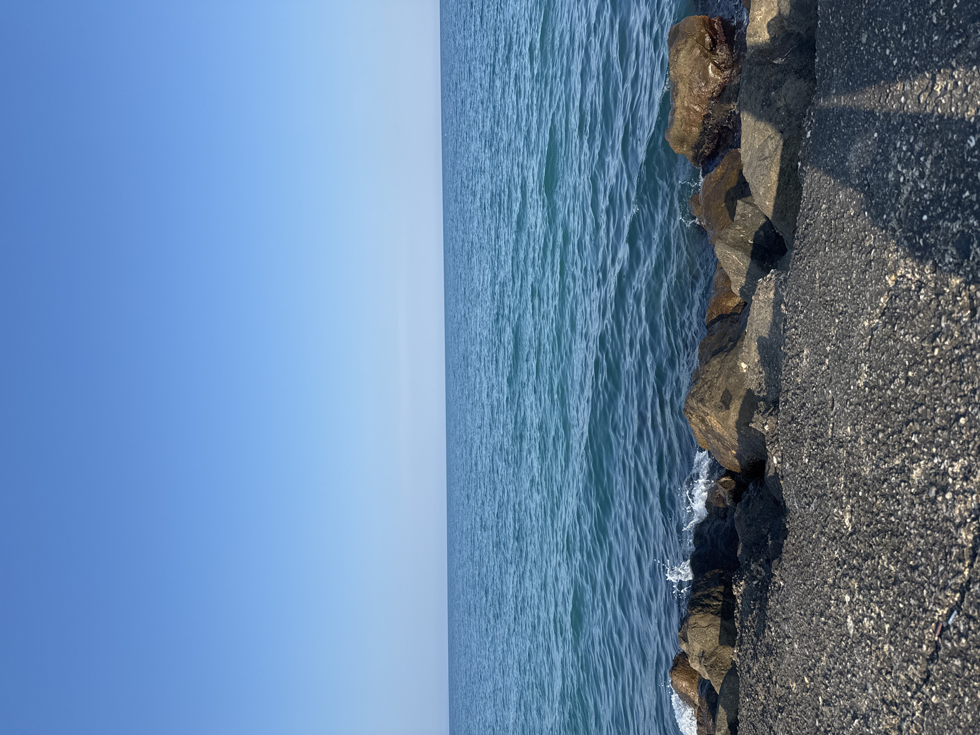 A wide-angle, eye-level shot captures a serene seascape in Pomorie, Bulgaria. The image is dominated by a vast expanse of clear blue sky, seamlessly meeting a tranquil turquoise sea. Gentle waves lap against a rocky shore in the foreground, composed of dark, coarse-grained asphalt or concrete, transitioning to larger, natural rocks of varying sizes and earthy tones. Some of the rocks are partially submerged in the water. The sunlight casts shadows across the shore, creating a play of light and dark on the textured surface. There are no people or animals visible in the frame. The overall impression is one of calm and natural beauty, characteristic of a seaside location on a clear day.