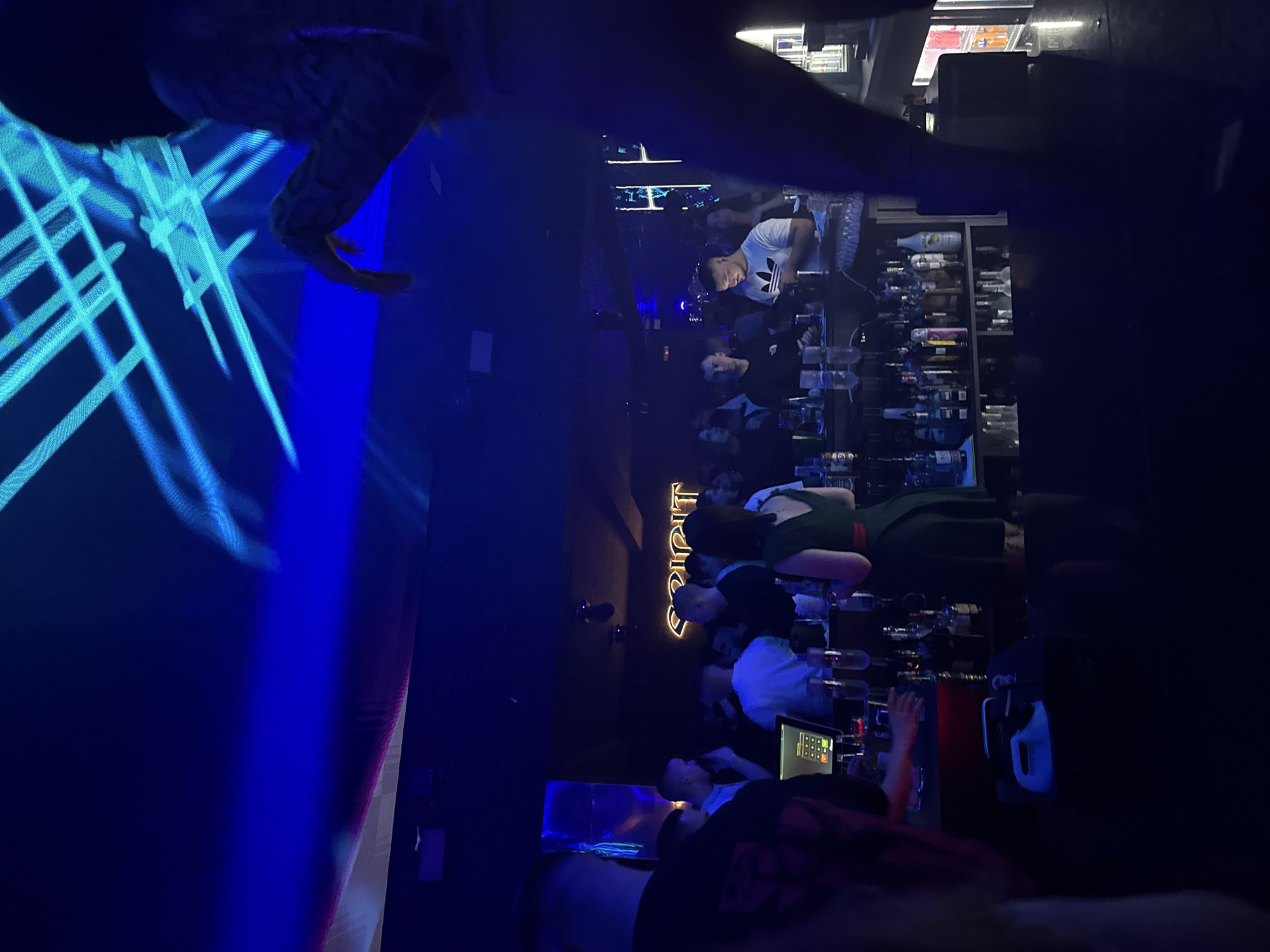 The image shows the interior of a dimly lit bar, possibly a nightclub, in Plovdiv, Bulgaria, given the neon sign that reads "SPIRIT" in Cyrillic. Several patrons are gathered at the bar counter, some standing and some seated on bar stools. A bartender is present behind the counter, possibly taking orders or preparing drinks. The bar is well-stocked with a variety of bottles, indicating an assortment of alcoholic beverages. A point-of-sale (POS) system is visible on the counter, suggesting ongoing transactions. The room is illuminated with blue and colored lights, potentially from a light display or DJ setup. A person with a snake is standing in front of a screen.