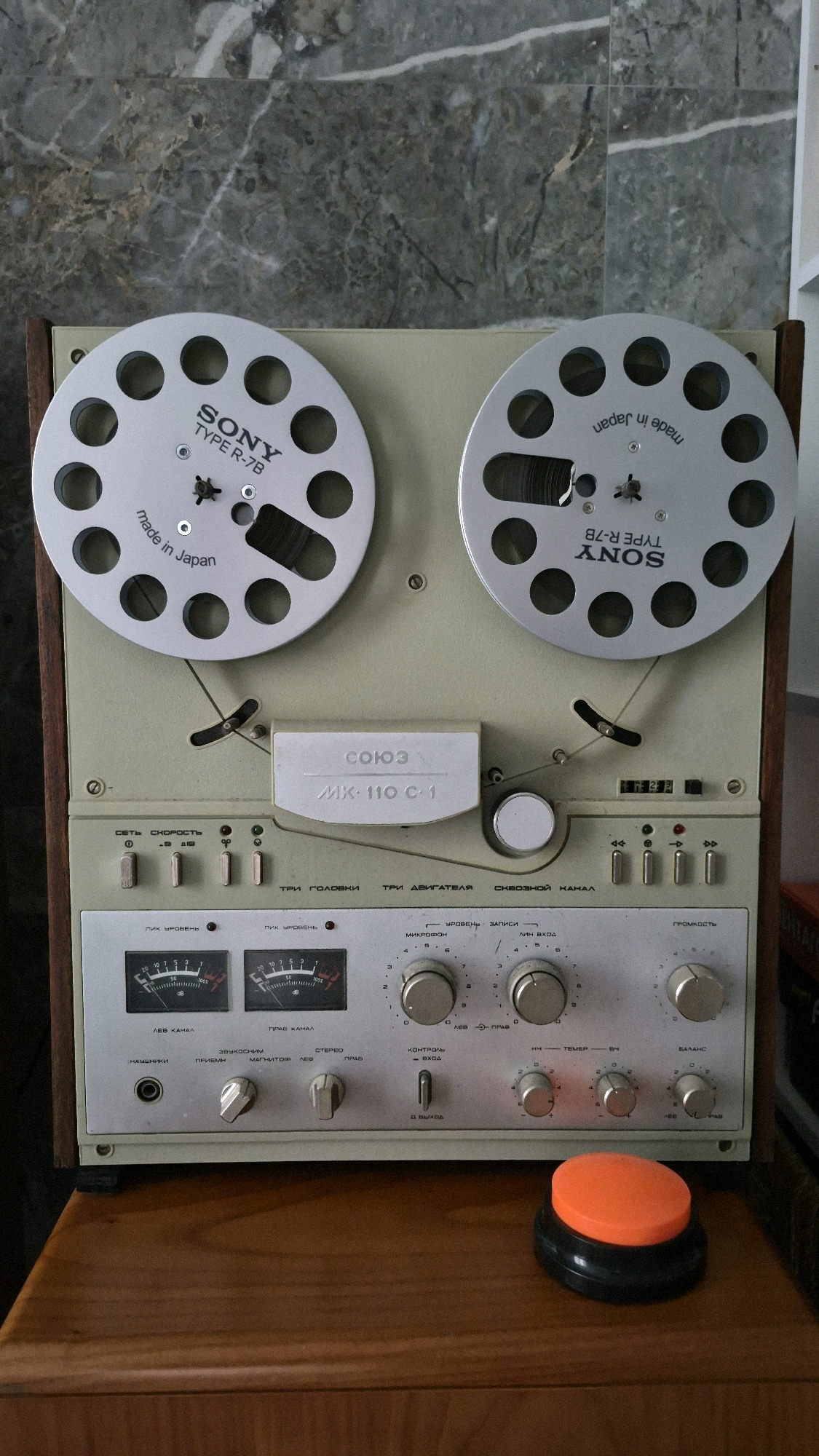 This is a close-up, eye-level shot of a vintage reel-to-reel tape recorder, likely a SONY Type R-7B, positioned on a wooden surface. The tape recorder is predominantly cream-colored with a wooden trim on its sides and features two large silver reels, one with "SONY TYPE R-7B" printed on it. The control panel is busy with various buttons, knobs, two VU meters, and labels in Russian. To the right of the tape recorder, a large orange and black button sits on the wooden surface. The background consists of a marbled wall. There are no people present in the image.