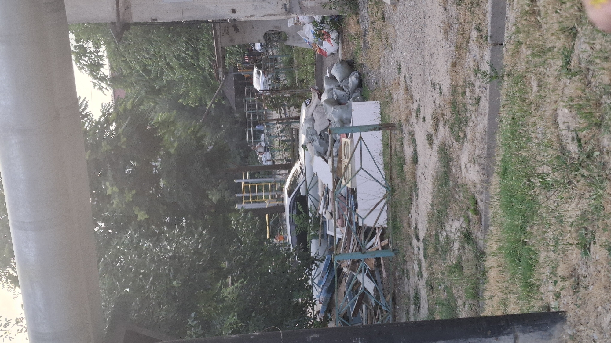 An outdoor scene in Shymkent, Kazakhstan, depicts a cluttered area with construction debris, including wooden planks, white boards, and sacks of material, piled near a white car. A metal fence separates the debris from a grassy area. In the background, trees and buildings are visible, along with a yellow and silver playground structure and other vehicles. A large pipe runs horizontally across the upper part of the frame.