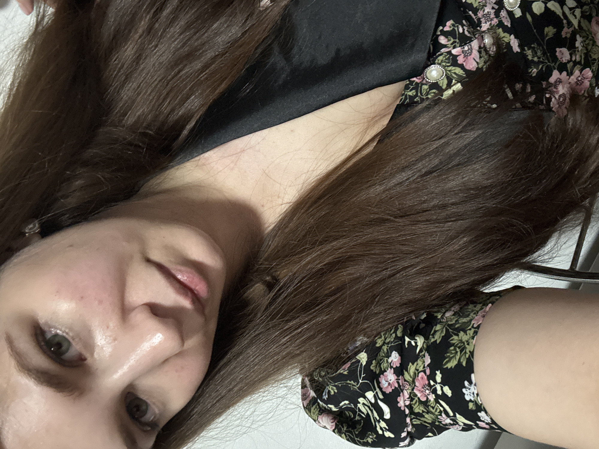 A close-up, head-and-shoulders shot of a young woman with long, dark brown hair, oriented vertically. She is looking down and slightly to her left. Her face is pale with visible pores and some redness on her cheeks. Her lips are slightly parted, and she has green eyes with dark eyeliner. She is wearing a black dress with a floral pattern of pink roses and green leaves. The dress has decorative pearl buttons. The background is mostly white, with some hints of what appears to be furniture. The lighting is bright, suggesting it could be daytime. The overall emotion conveyed is casual and relaxed. There is no visible text in the image.