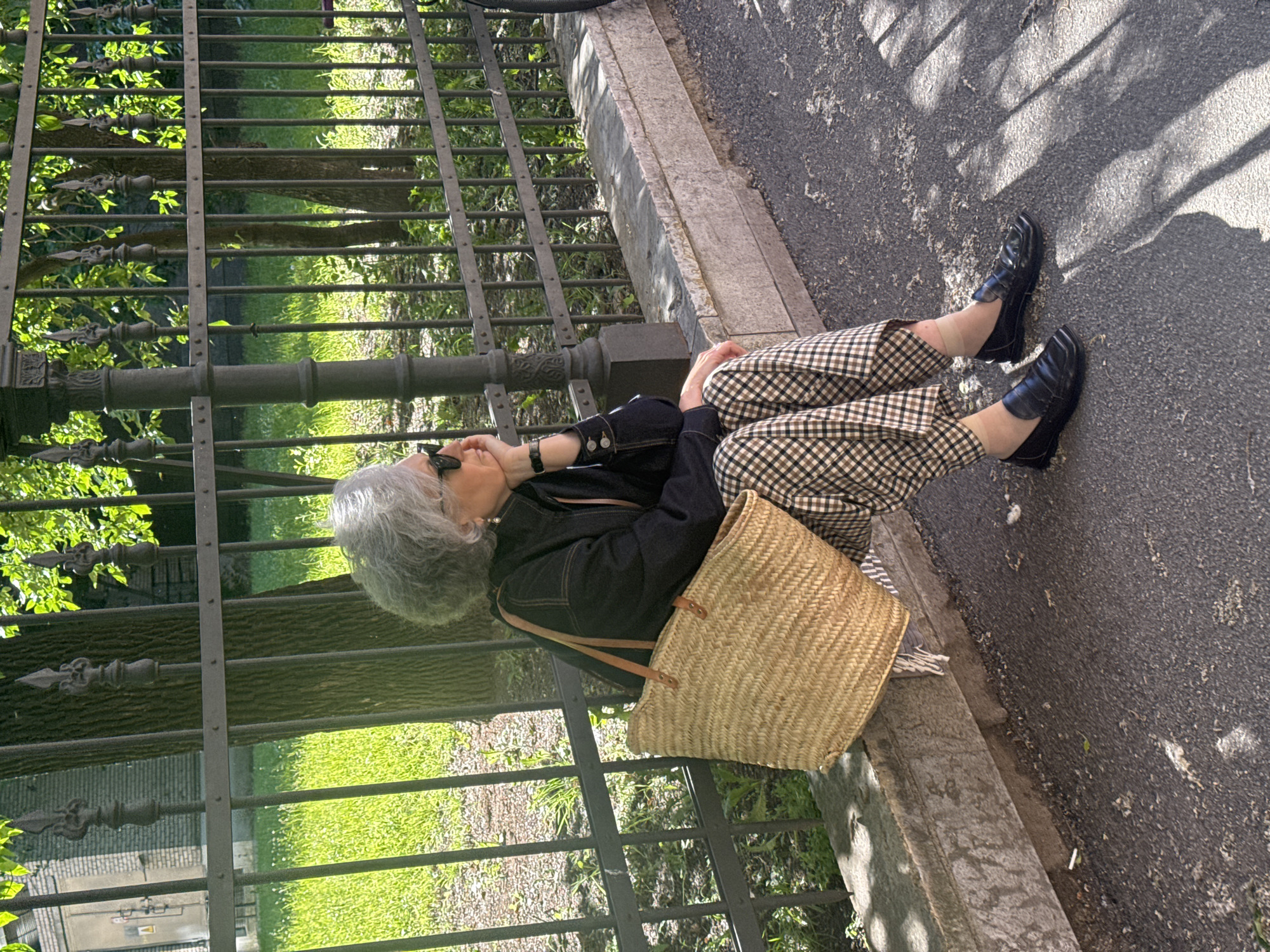 A woman with gray hair is sitting on a concrete ledge next to a black metal fence. She is wearing a dark denim jacket, plaid pants, and black loafers. A woven straw bag with leather straps rests beside her on the ledge. She is holding a phone to her ear and appears to be talking. The scene is outdoors on a sunny day, with dappled sunlight casting shadows on the asphalt path. Green grass and trees are visible behind the fence. The image was taken in Saint Petersburg, Russia.