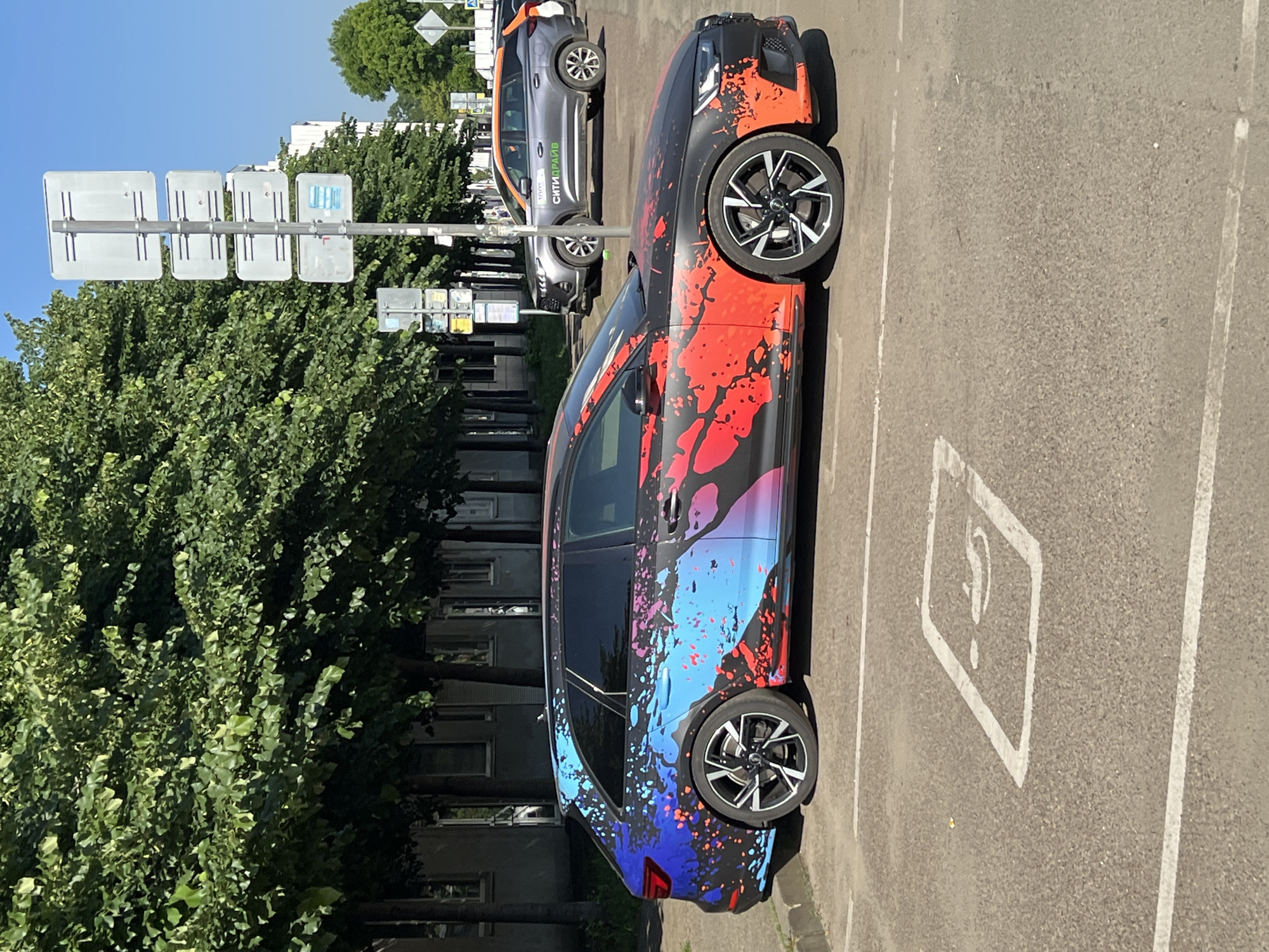 The image presents an urban street scene, likely a parking area in Krasnodar, Russia, under clear blue skies and bright daylight conditions.
The central subject is a black Audi station wagon, specifically an Avant model, parked diagonally in the foreground. The vehicle is distinctive due to its vibrant, custom full-body wrap featuring an abstract paint splatter design. This livery transitions from shades of blue and purple on the rear half of the car to red and orange on the front half, applied over the black base. The car is equipped with multi-spoke black and silver alloy wheels.
The ground consists of asphalt pavement, marked with faded white parking lines. In front of the Audi's left front wheel, a white square ground marking contains an indistinct white symbol, possibly indicating a specific parking type, partially obscured by the vehicle.
In the mid-ground, behind and to the right of the Audi, a grey crossover SUV is parked. This vehicle is identified as a "СИТИДРАЙВ" (CityDrive) car-sharing service vehicle by green text on its side and orange accents on its roof rails and rear.
The background on the left is dominated by a dense row of mature deciduous trees with vibrant green foliage, suggesting a street lining or park area. Partially visible behind these trees are multi-story buildings with light-colored facades and numerous windows. To the right of the trees and behind the parked vehicles, a tall metal pole supports a stack of four large, rectangular white-backed street signs facing away from the camera. A smaller blue sign is discernible on the lowest of these. Further down the street, another pole with additional signs and a pedestrian crossing sign are visible on the far right.
No individuals are present in the visible frame. The strong sunlight indicates fair weather, casting defined shadows on the pavement. The overall setting is consistent with a typical urban street and parking environment found in a Russian city.