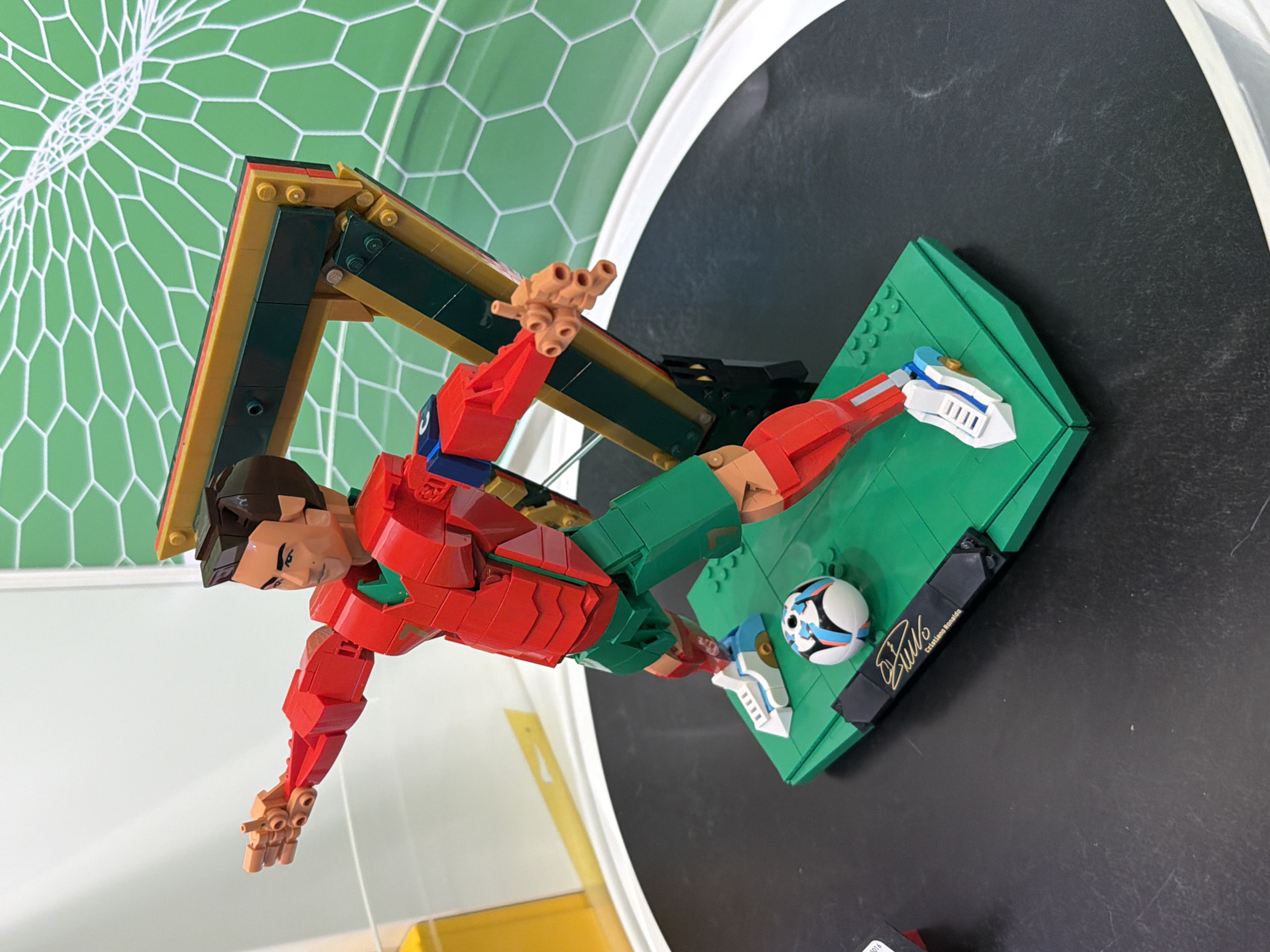 The image shows a Lego model of Cristiano Ronaldo, positioned as if he is leaping to score a goal. He is in a red and green soccer uniform, with a soccer ball at his feet on a green Lego base. A Lego soccer goal is behind him, with a green net design on the wall in the background. The model is displayed on a black surface, possibly a table, within a white, rounded enclosure. The setting is a museum, specifically the Museo del Automóvil Puebla in Puebla, Mexico.
