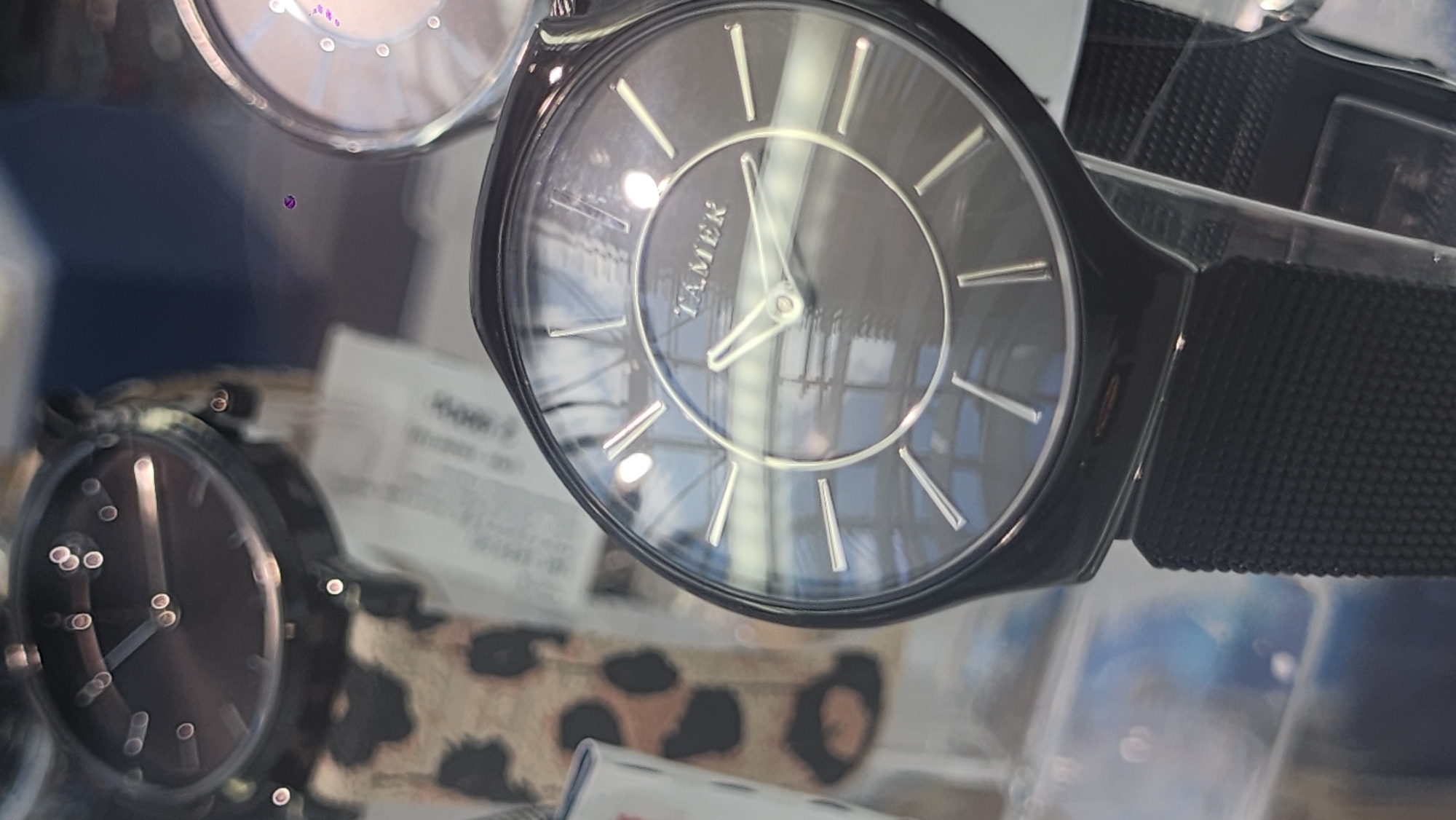 A close-up, vertically oriented, eye-level shot shows two analog watches with dark faces and silver hands and markers. The watch in the foreground, on the right side of the frame, is a TAMER brand watch with a mesh metal band. It has a dark face with silver hour markers and hands. The watch in the background, to the left, has a leopard-print band. The watches appear to be displayed on a shelf or in a case, as suggested by the blurred background and reflections. The lighting is somewhat dim, creating reflections on the watch faces.