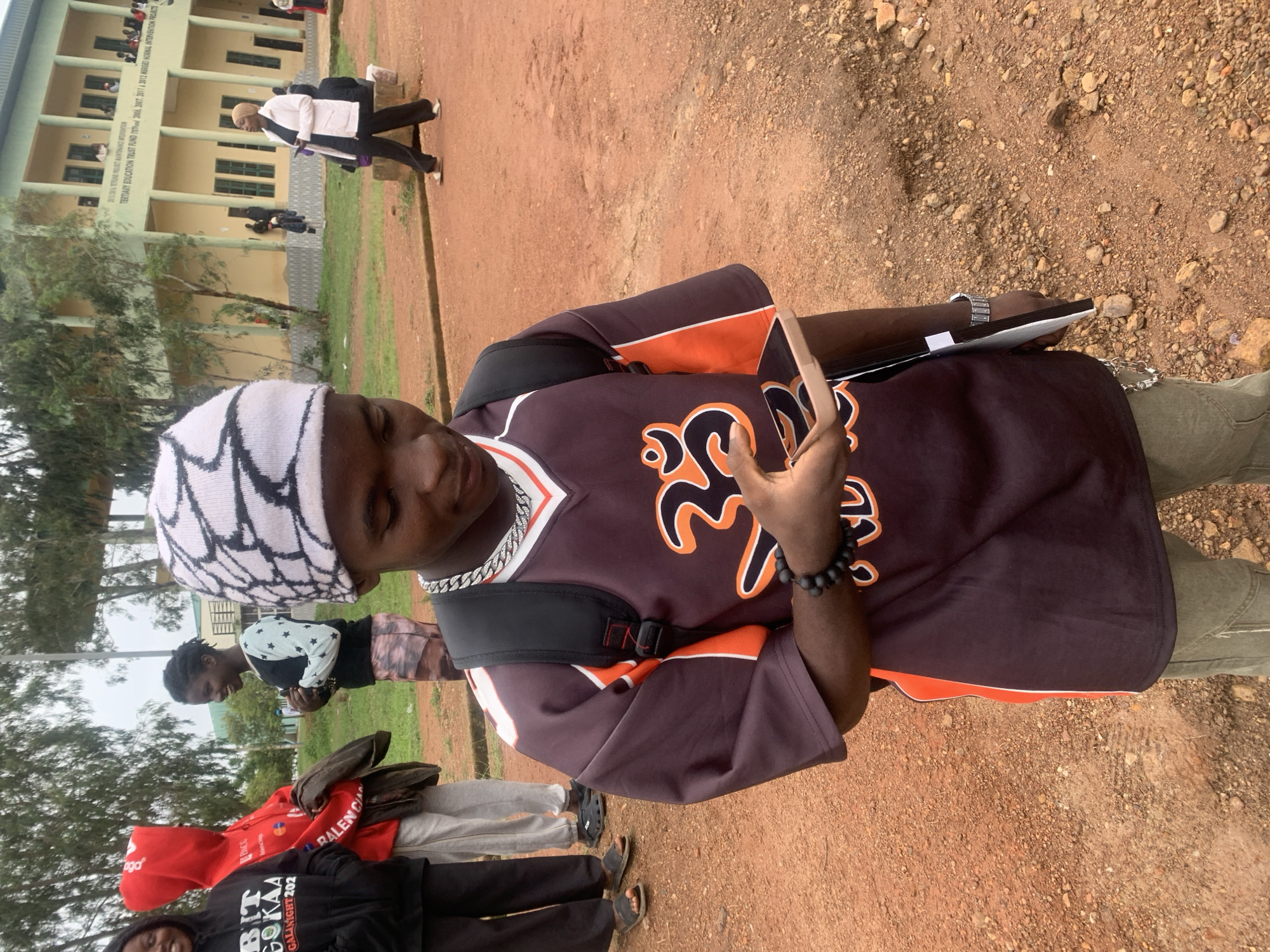 In Durumi, Nigeria, a young person stands outdoors, looking down at a smartphone. They wear a dark jersey with orange accents and a white beanie with a black pattern. Around their neck is a chunky silver chain, and on their wrist, a beaded bracelet. To the left, three other young people are visible. In the background, a building with multiple stories and windows stands under a cloudy sky, surrounded by trees and a dirt path.
