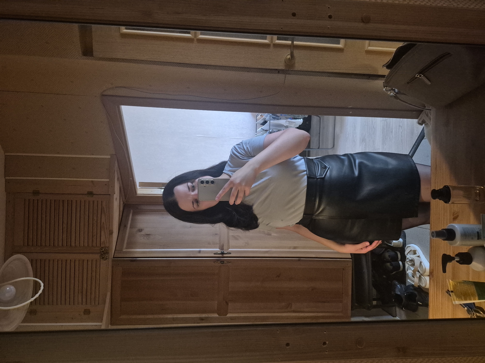 The image shows a woman in a light blue t-shirt and a black faux leather skirt, taking a mirror selfie in a room with wooden walls and cabinets. She is standing in front of a large mirror that reflects her and a portion of the room. A light fixture with a circular shade hangs from the ceiling above the cabinets. In the reflection, a doorway is visible, leading to another room. Below the cabinets, a shelf holds several pairs of shoes, and a few cosmetic bottles and a purse are on a wooden surface in the foreground. The overall setting appears to be a domestic interior, likely a bedroom or dressing area. The specific location in Saint Petersburg, Russia, cannot be determined from the visual information provided.
