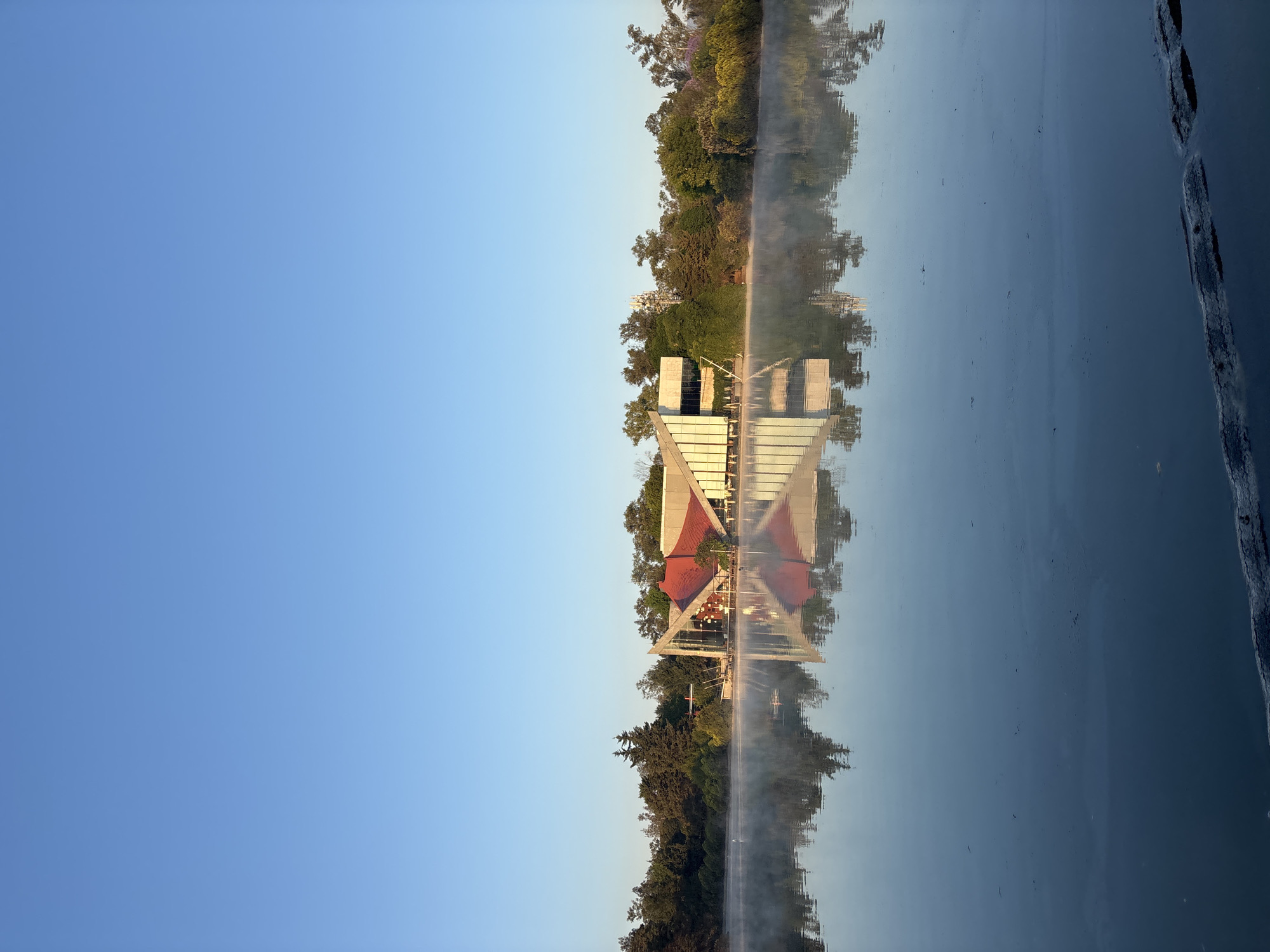 The image shows a body of water reflecting a building and trees. The building has a red roof and a triangular shape. The water is calm and reflects the blue sky above. There is some mist rising from the water. The trees are green and lush. The sky is clear and blue. The image is taken in an outdoor setting. The city is Unknown, Unknown.